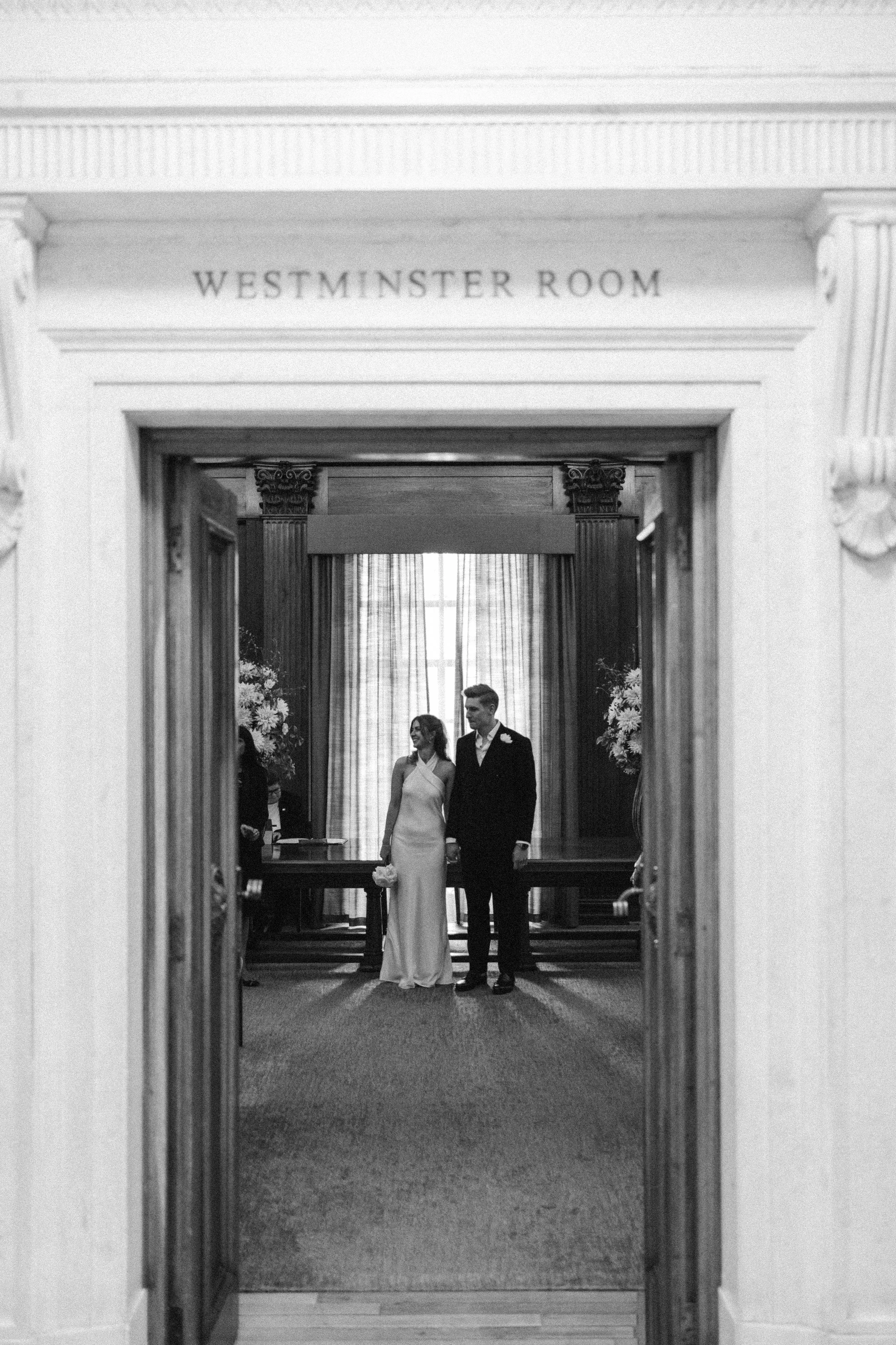 A wedding couple standing at the entrance of the Westminster Room in a formal setting, with the bride in a white dress and the groom in a dark suit, surrounded by floral arrangements and draped curtains.