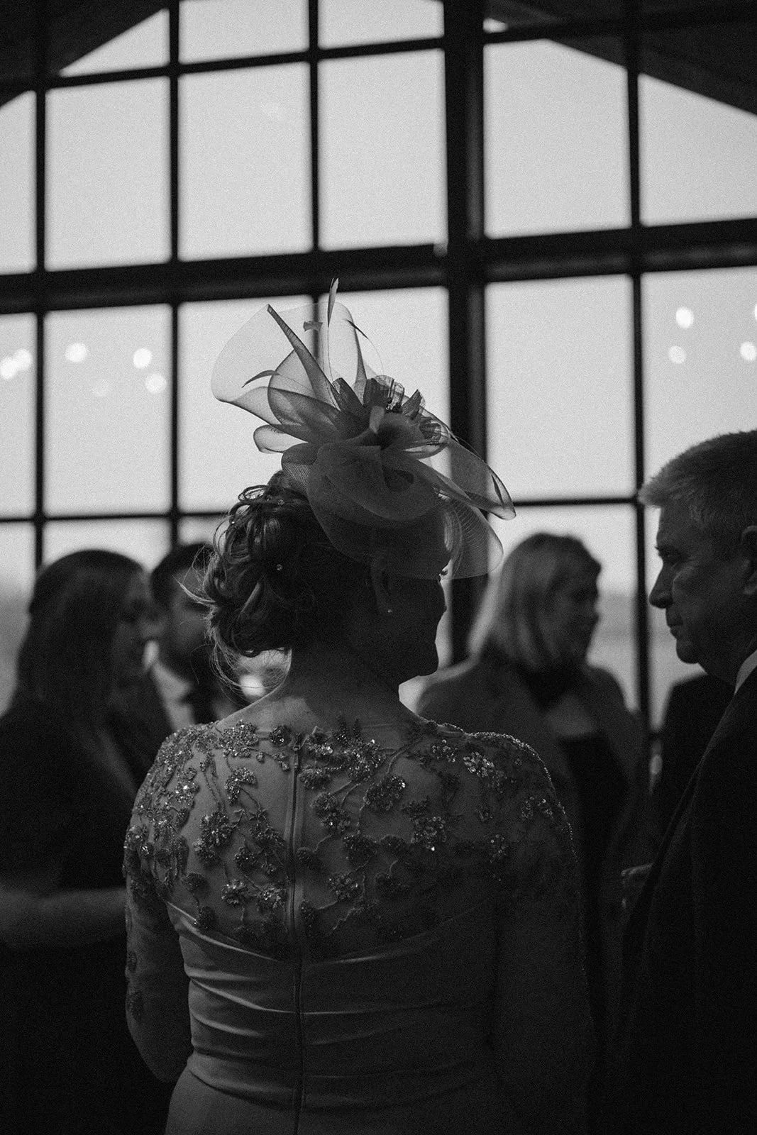 Silhouette of a woman in an embellished dress with a large fascinator on her head, standing indoors with a large window and several people in the background.