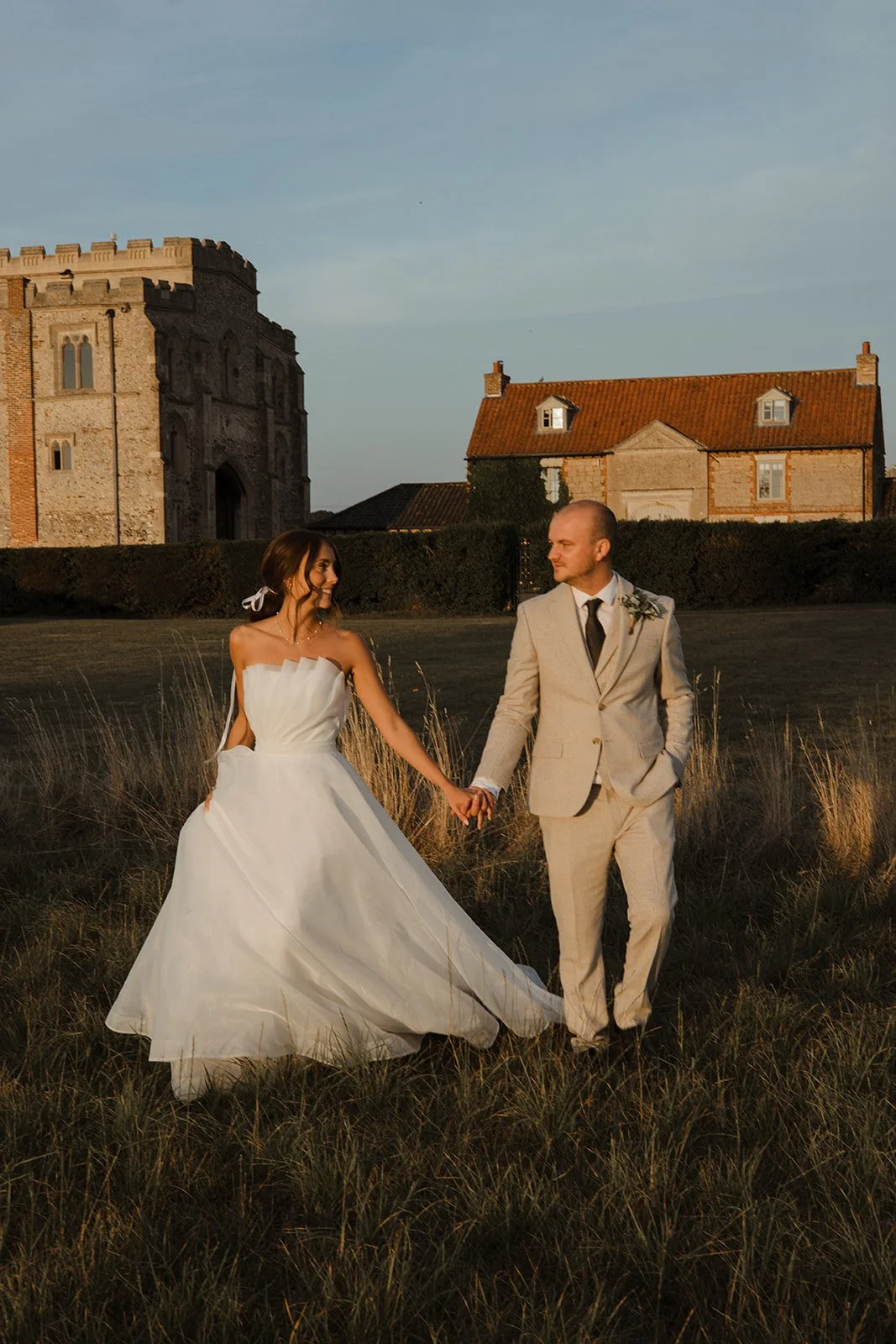 A bride and groom holding hands in a field during sunset, with historic buildings in the background.