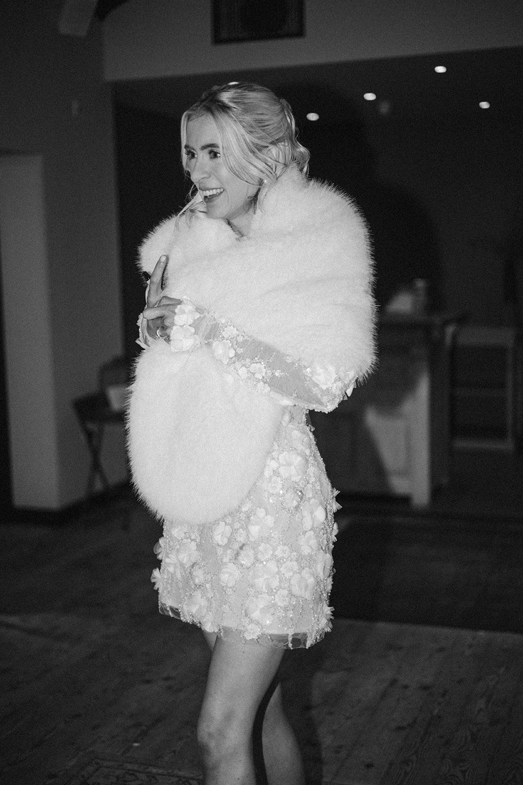 A smiling woman in a sparkly dress and a fluffy shawl, holding a fuzzy clutch purse, standing indoors with a wooden floor and dark background.