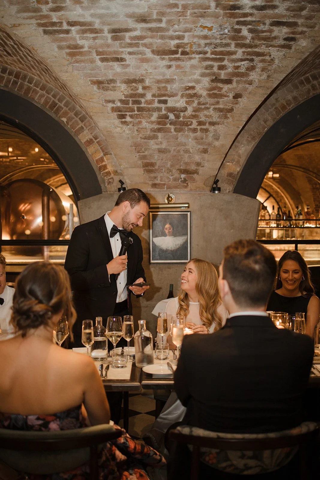 A man in a black tuxedo and bow tie is giving a speech or toast at a dinner event, holding a microphone in one hand and a phone in the other. He is smiling and looking down at a woman with long blonde hair, who is smiling back at him. The setting is 