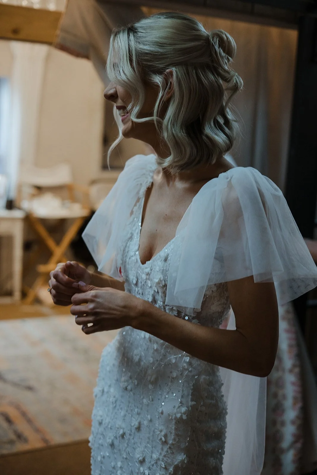 A woman in a wedding dress with puffed tulle shoulders, smiling and clasping her hands, standing in a rustic room.