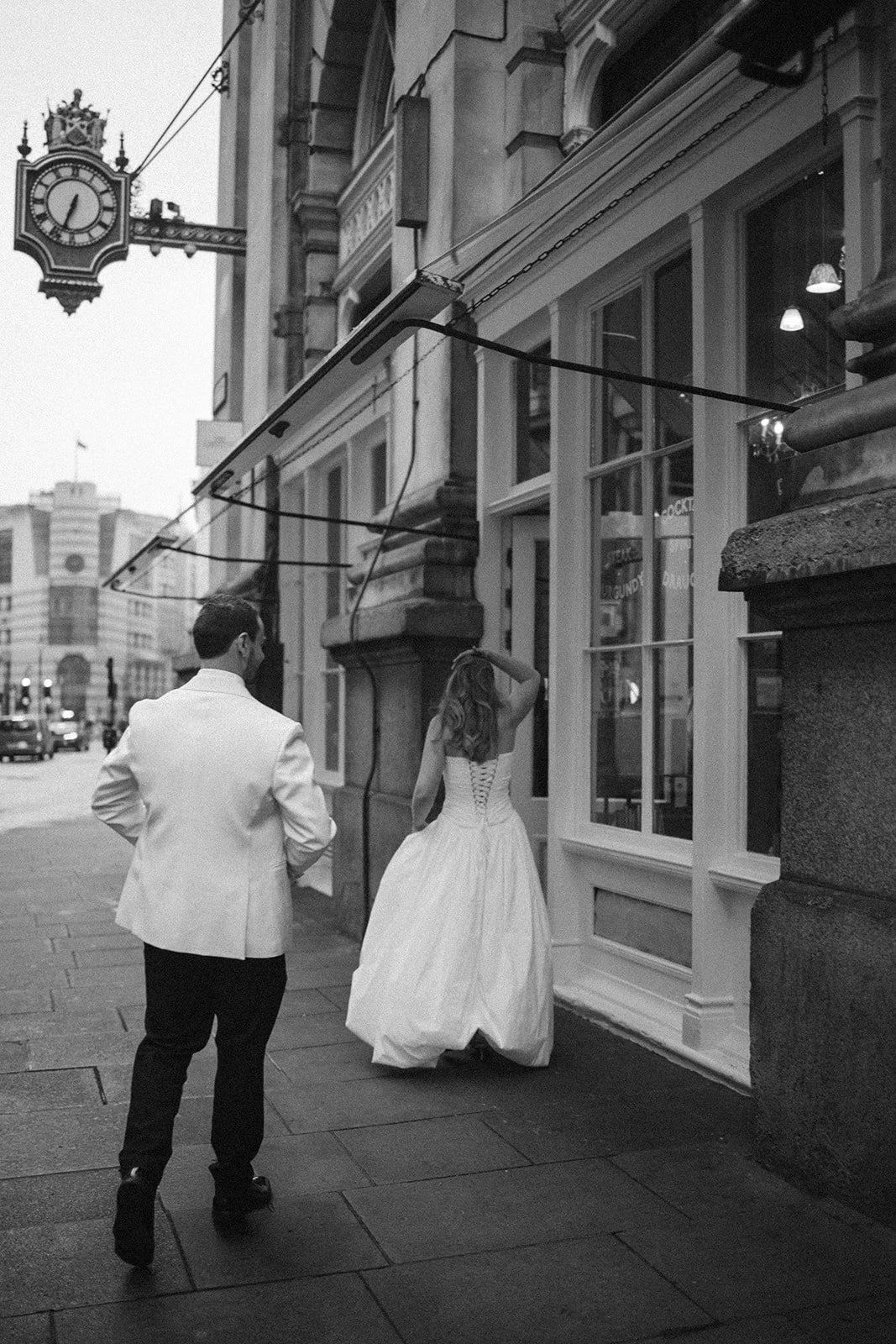 A man and a woman in vintage formal attire walking down a city sidewalk at night, with a street clock and buildings in the background.