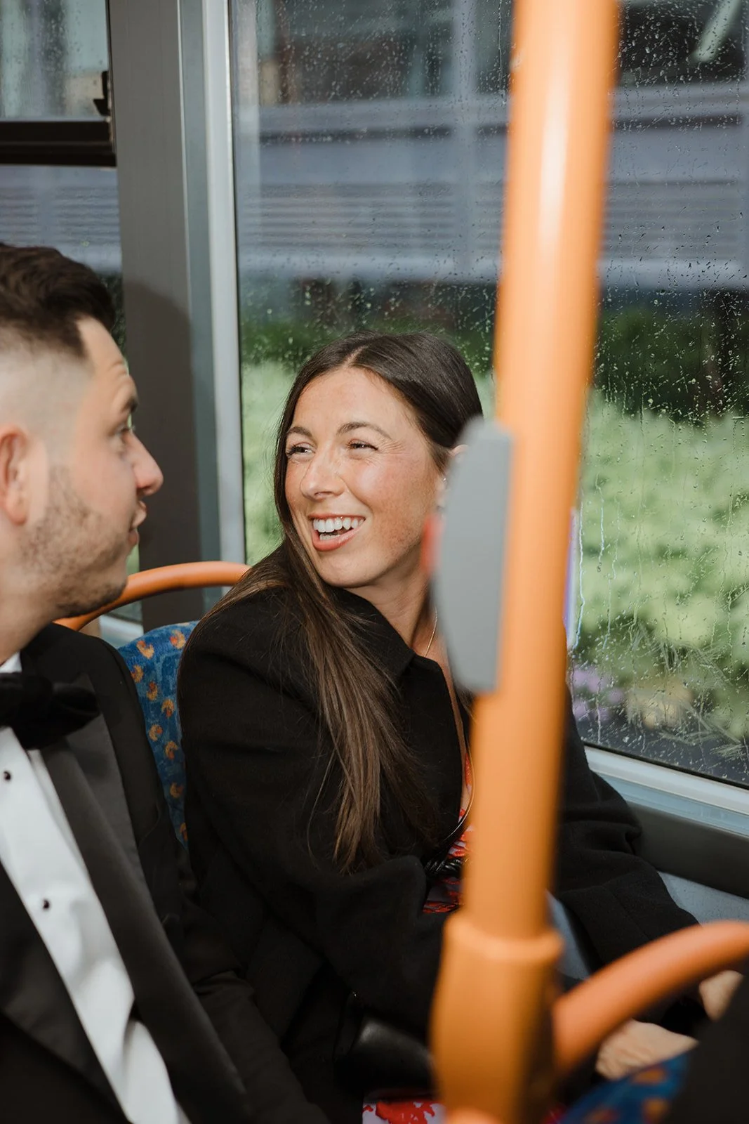 A woman and man sitting on a bus, the woman is smiling and looking at the man, with a rain-covered window behind them.
