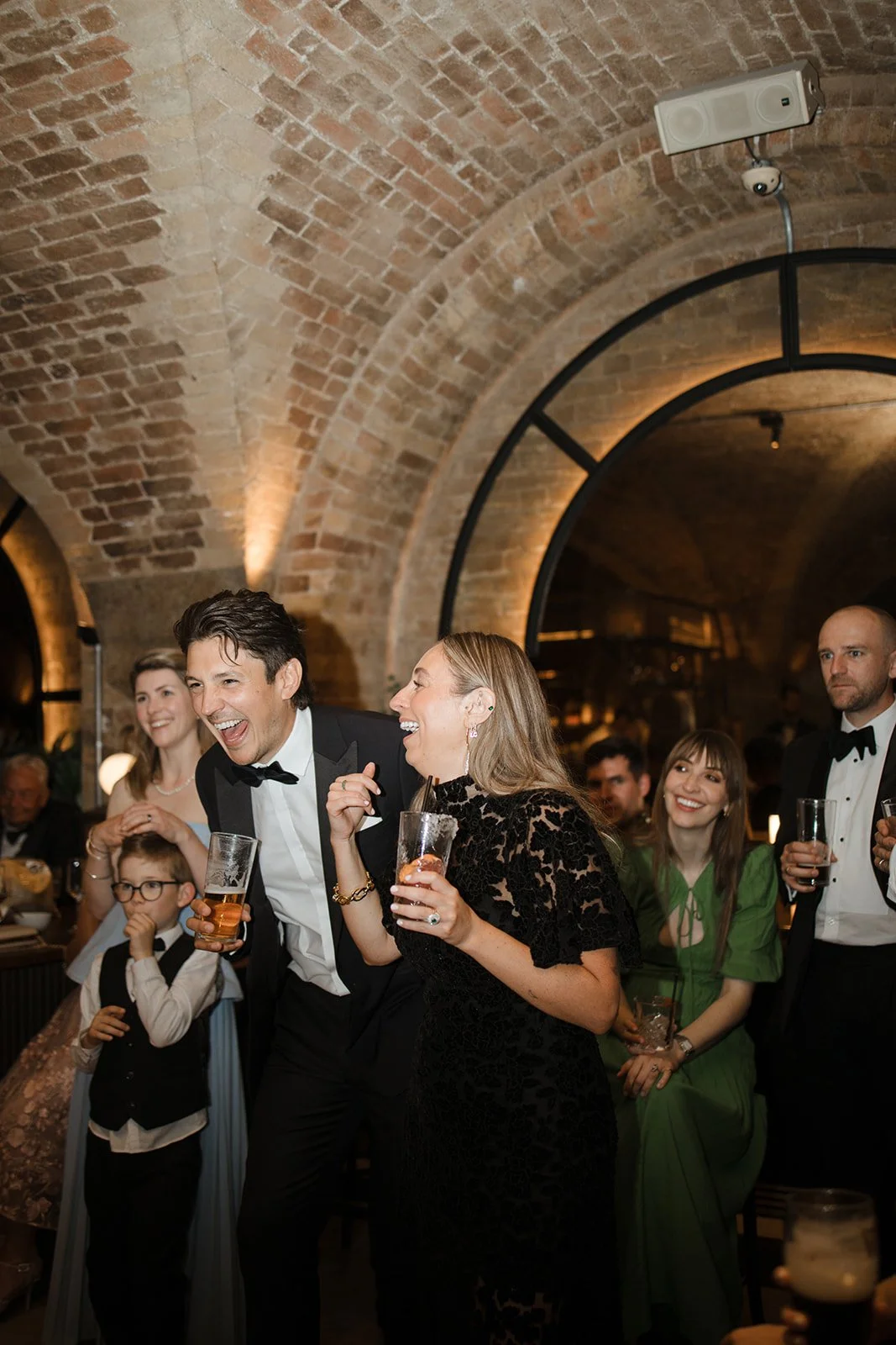People dressed in formal attire celebrating and enjoying drinks at a party in a brick-arched venue.