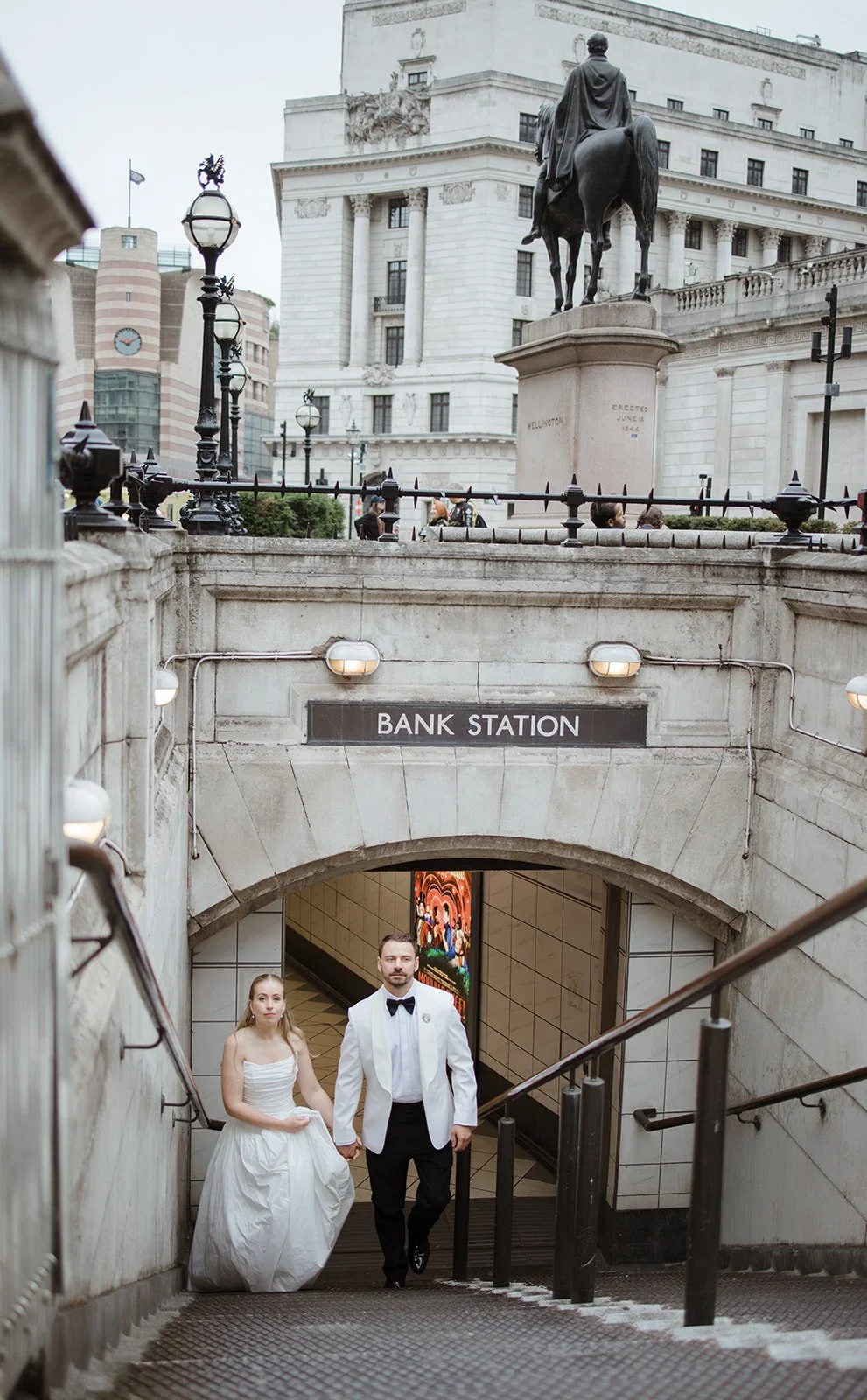 A couple dressed in wedding attire walking up the stairs at a subway entrance labeled 'Bank Station'. The man is wearing a tuxedo with a bow tie, and the woman is wearing a white wedding dress. An equestrian statue and classical building are visible 