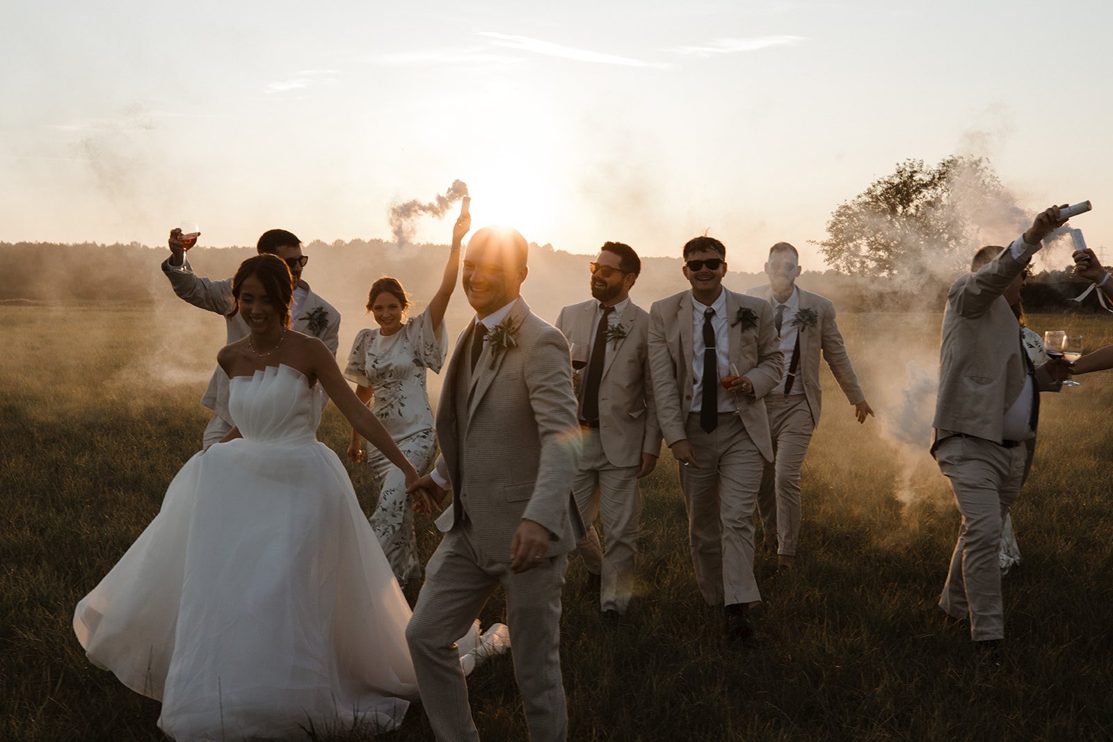 A joyful wedding celebration outdoors at sunset with the bride and groom holding hands, surrounded by friends in light-colored suits and floral dresses. Smoke and sparklers add to the festive atmosphere.