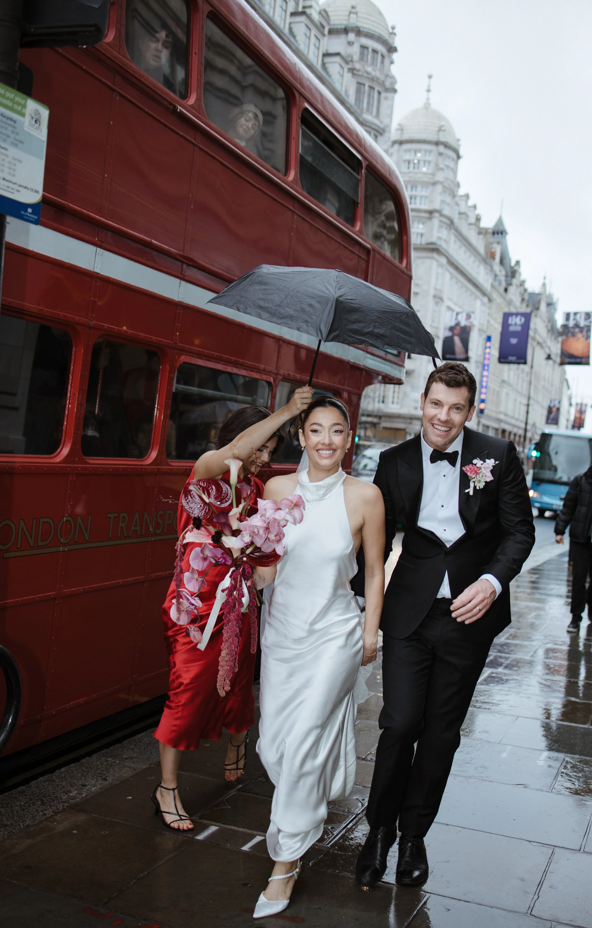 A bride and groom walking on a wet city sidewalk after a wedding, with a woman holding an umbrella over the bride. A red double-decker bus and historic buildings are in the background.