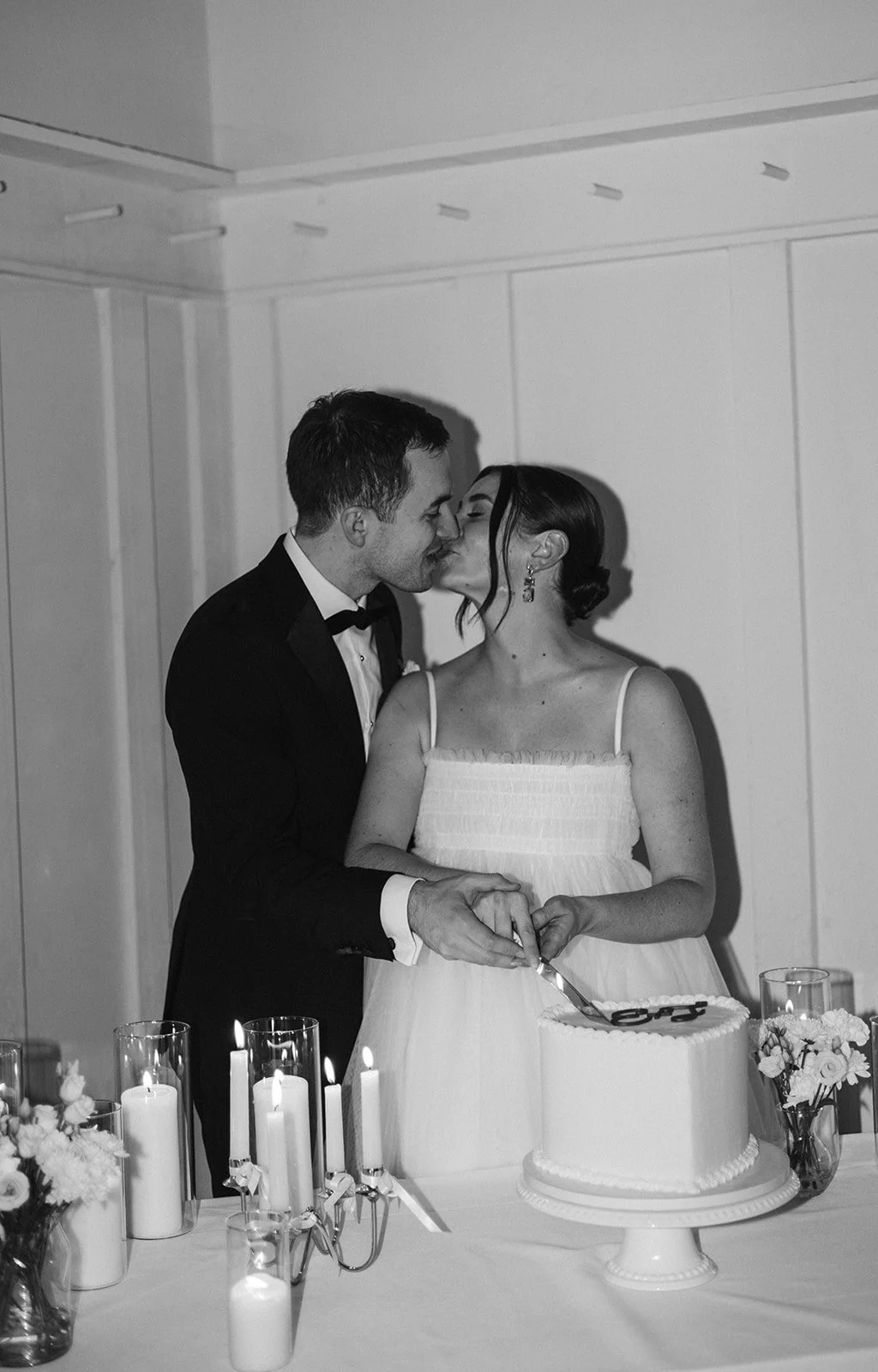 A black-and-white photo of a bride and groom kissing during their wedding reception, with the bride holding a cake knife over a wedding cake, candles, and floral arrangements on the table.