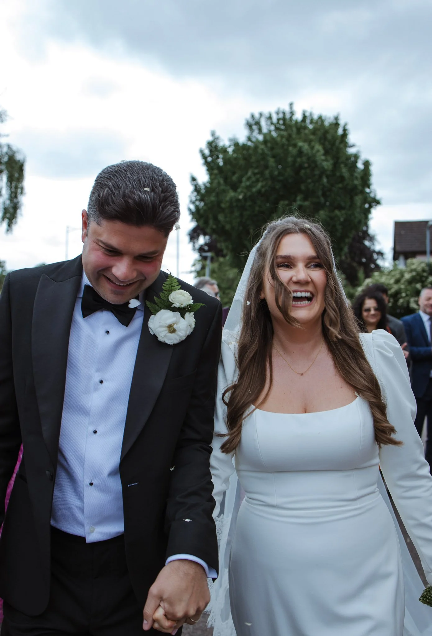 A smiling bride and groom holding hands outdoors during a wedding ceremony, with guests in the background.