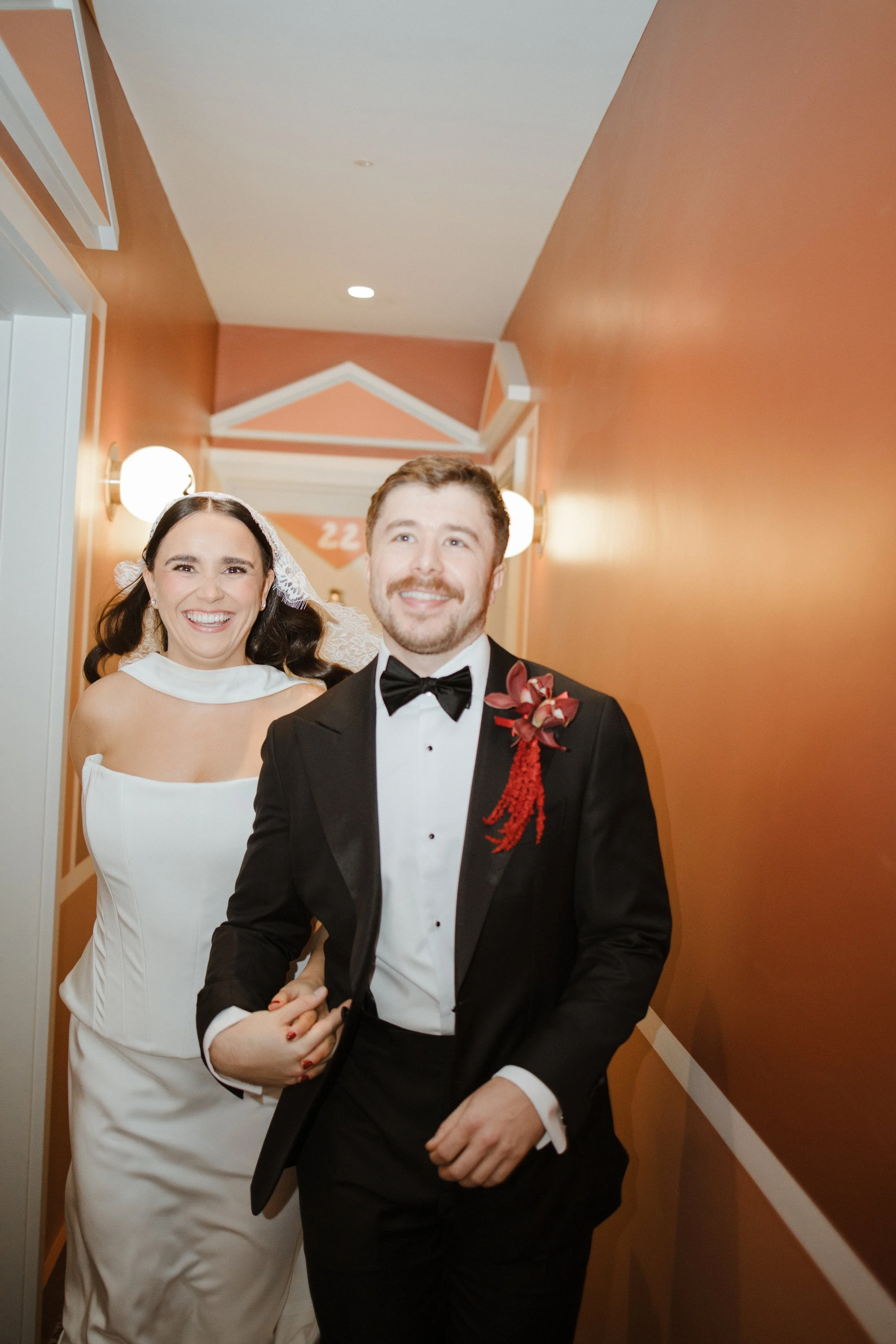 A newlywed couple walking down a hallway, smiling at the camera. The bride is wearing a white wedding dress and a veil, and the groom is dressed in a black tuxedo with a bow tie and a red flower boutonniere.
