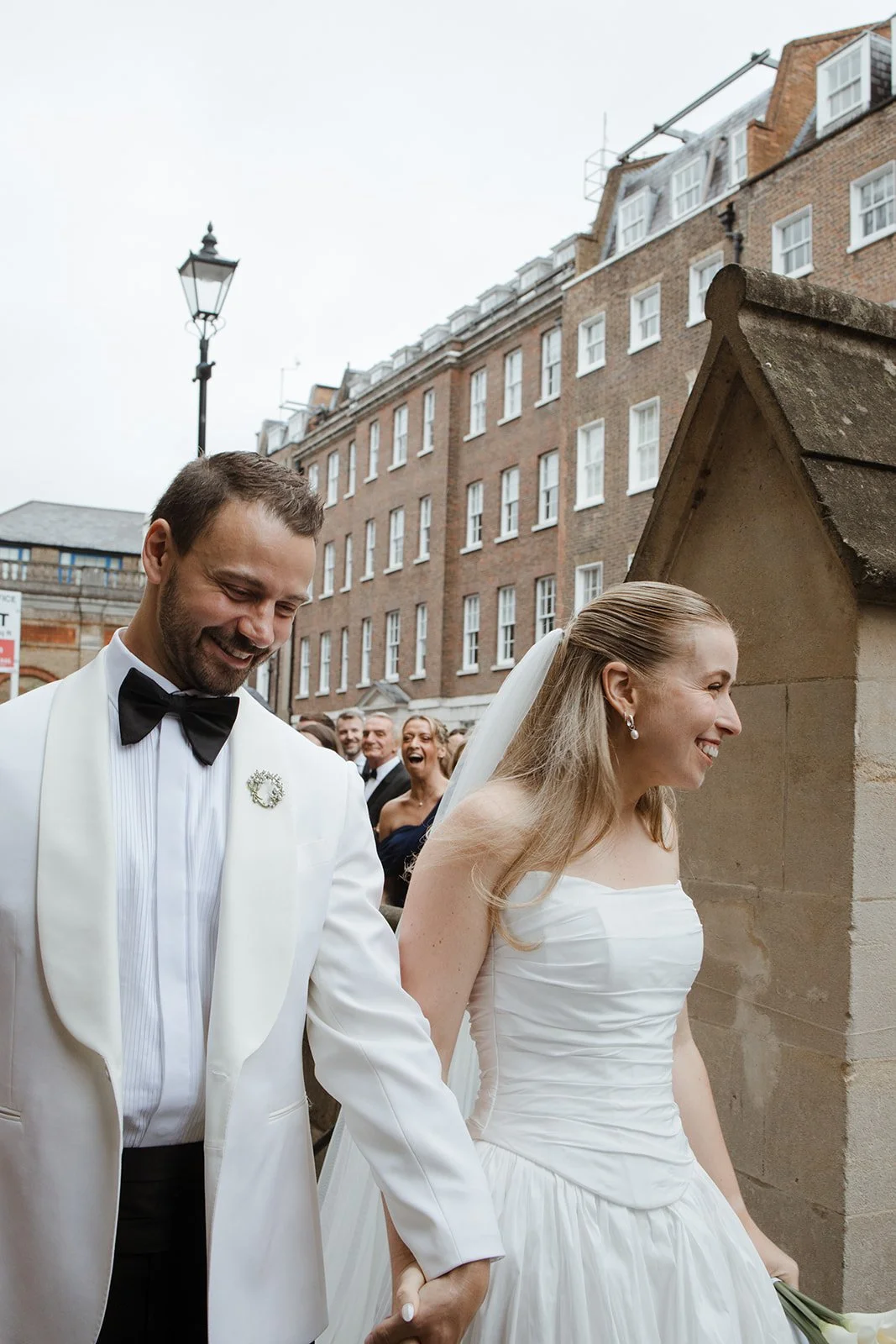 A happy couple dressed in wedding attire holding hands and smiling, surrounded by friends outside a church or historic building with brick architecture.