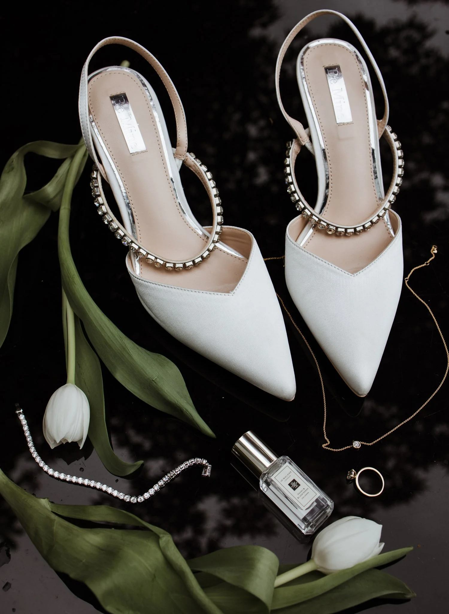 Pair of white pointed-toe heels with embellished ankle straps, surrounded by white tulips, jewelry, and a perfume bottle on a black surface.