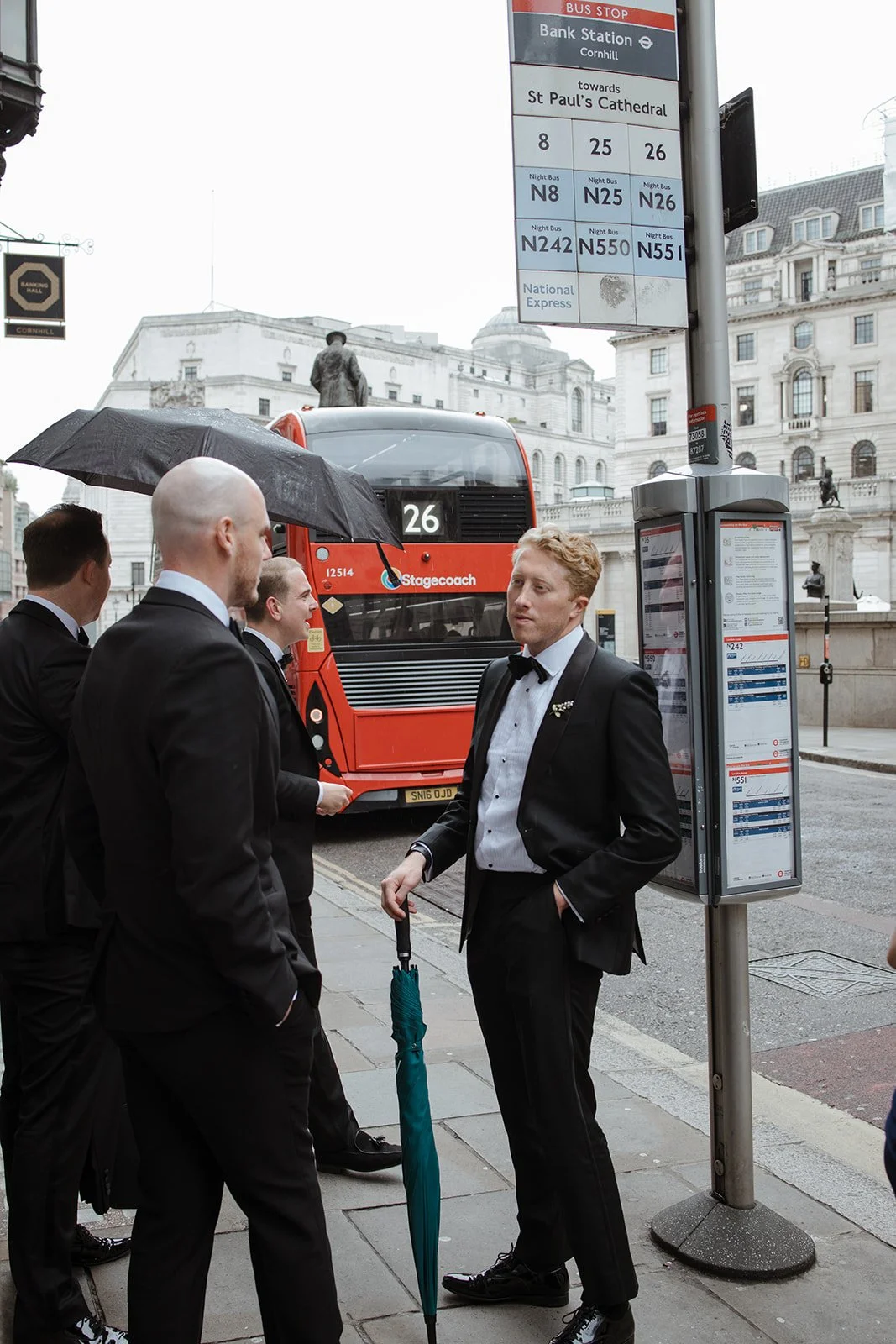 Group of men in tuxedos standing near a bus stop on a city street, one man holding an umbrella, in rainy weather, with historic buildings in the background.