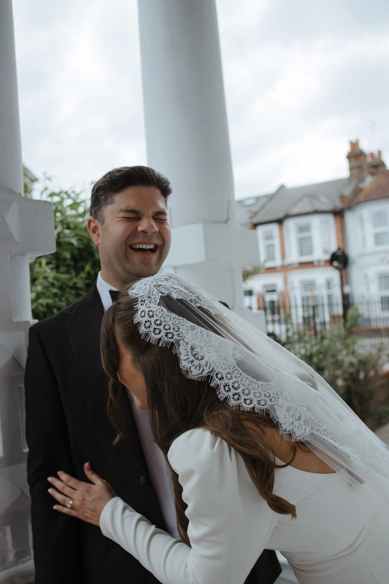 A joyful bride and groom sharing a moment, with the bride leaning in and both smiling, outdoors near a white column.