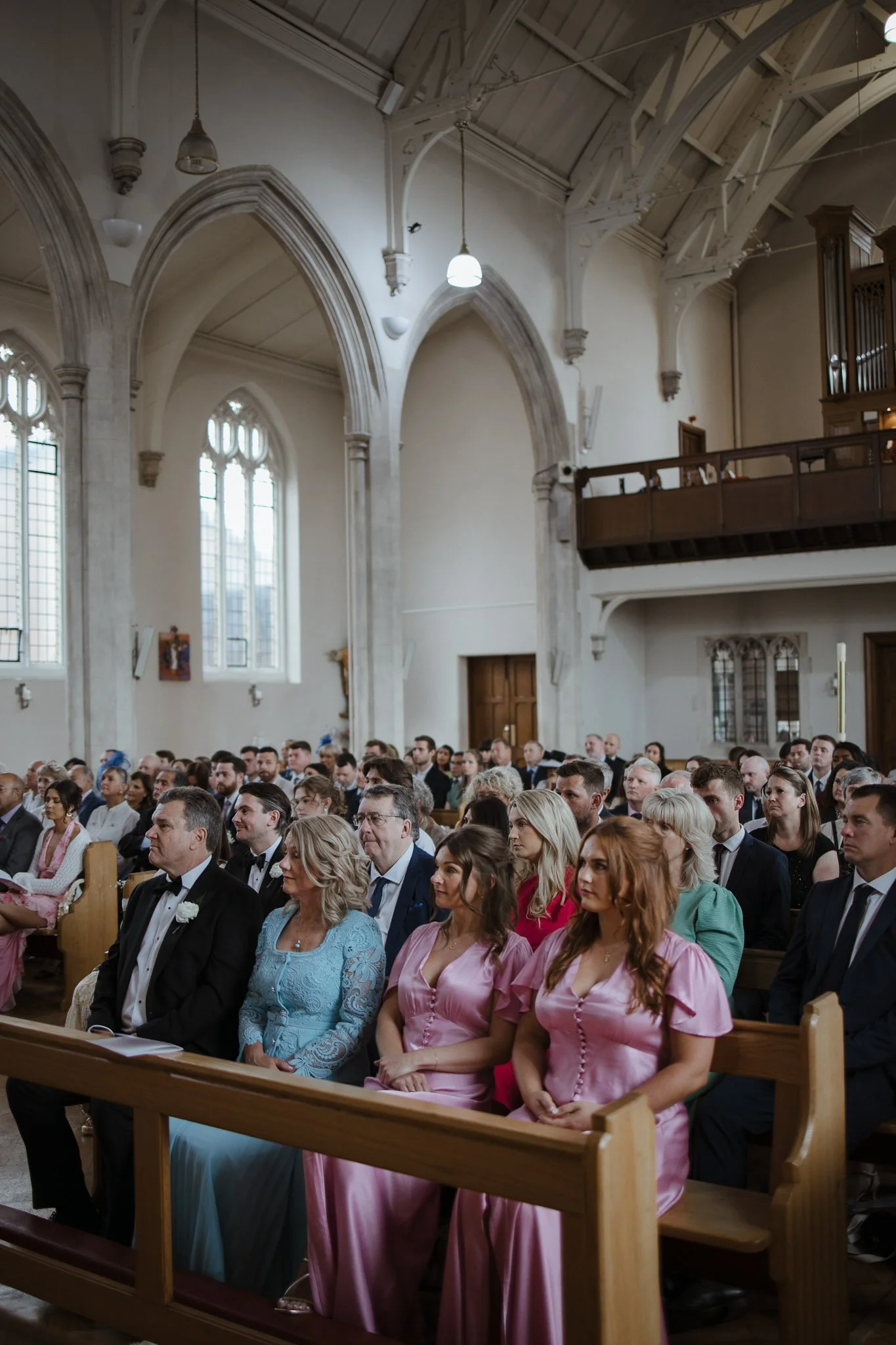 People seated in pews during a wedding ceremony inside a church with high vaulted ceilings and tall stained glass windows.