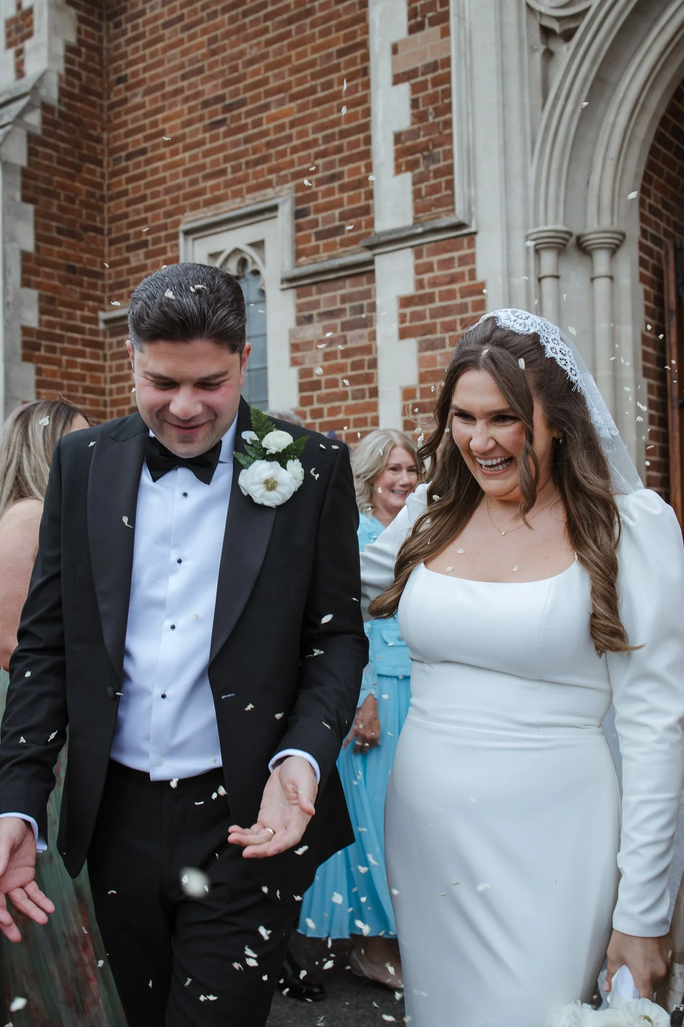 Bride and groom walking outside a brick church, smiling and surrounded by guests, as confetti falls around them.