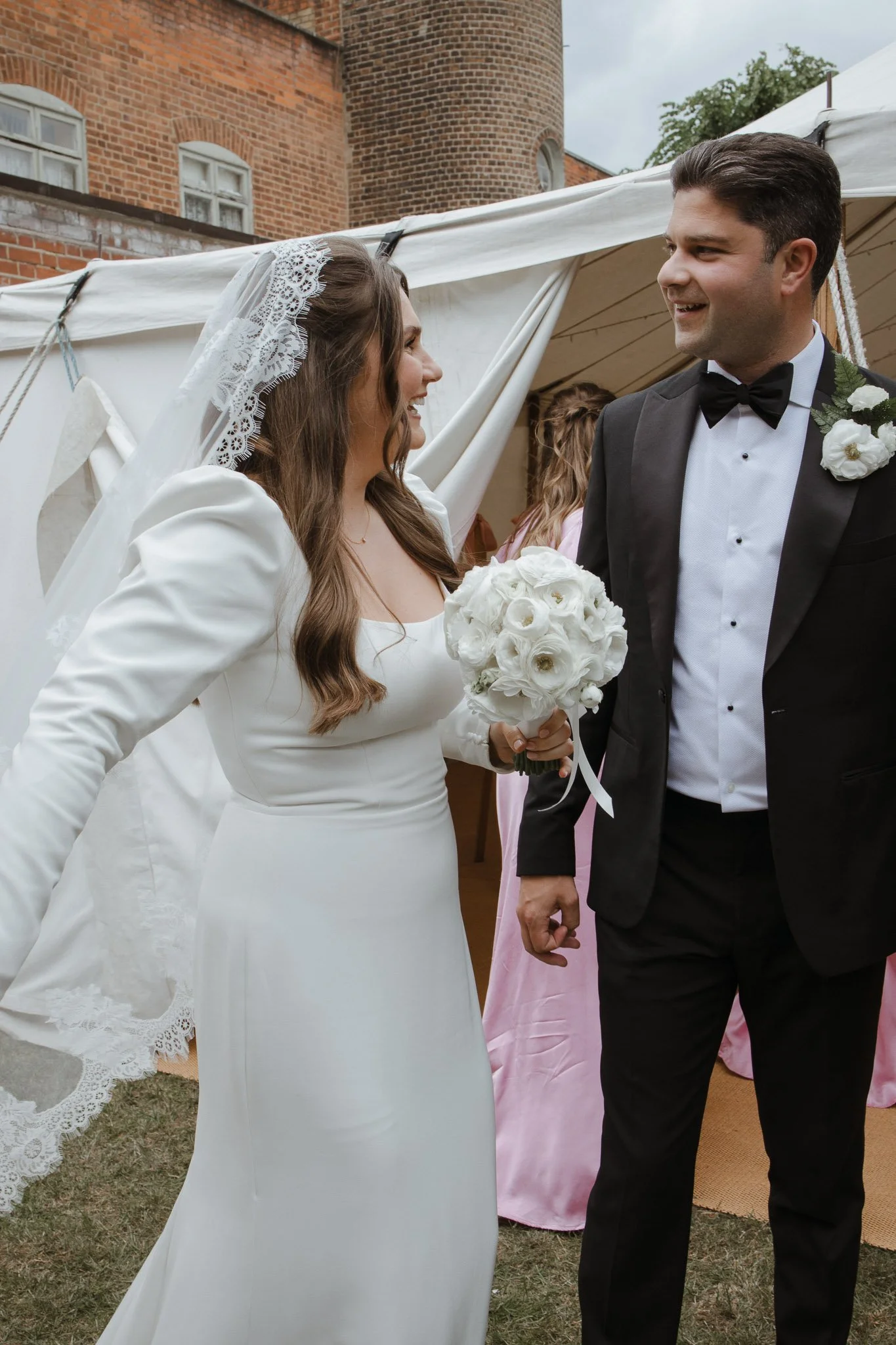 A bride and groom talking at their wedding outdoors, with the bride holding a bouquet of white roses, both smiling and dressed in formal wedding attire.