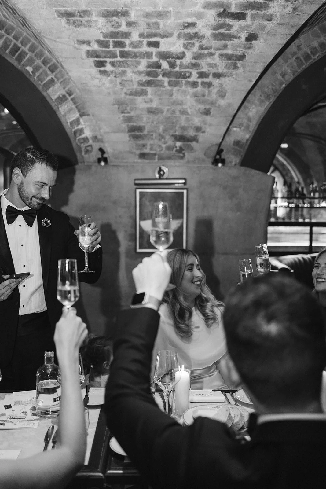 Black and white photo of people celebrating at a dinner party in a restaurant or bar with brick arches on the ceiling. The man in a tuxedo is holding a glass of champagne and smiling, while women are raising glasses and toasting, some seated at the t