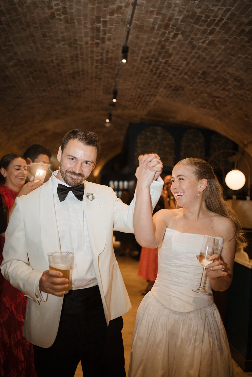 A man and woman dancing at a celebration, holding hands, with other guests in the background. The man is wearing a white tuxedo jacket with a black bow tie, and the woman is in a strapless white dress. The setting is an arched brick-ceiling venue.