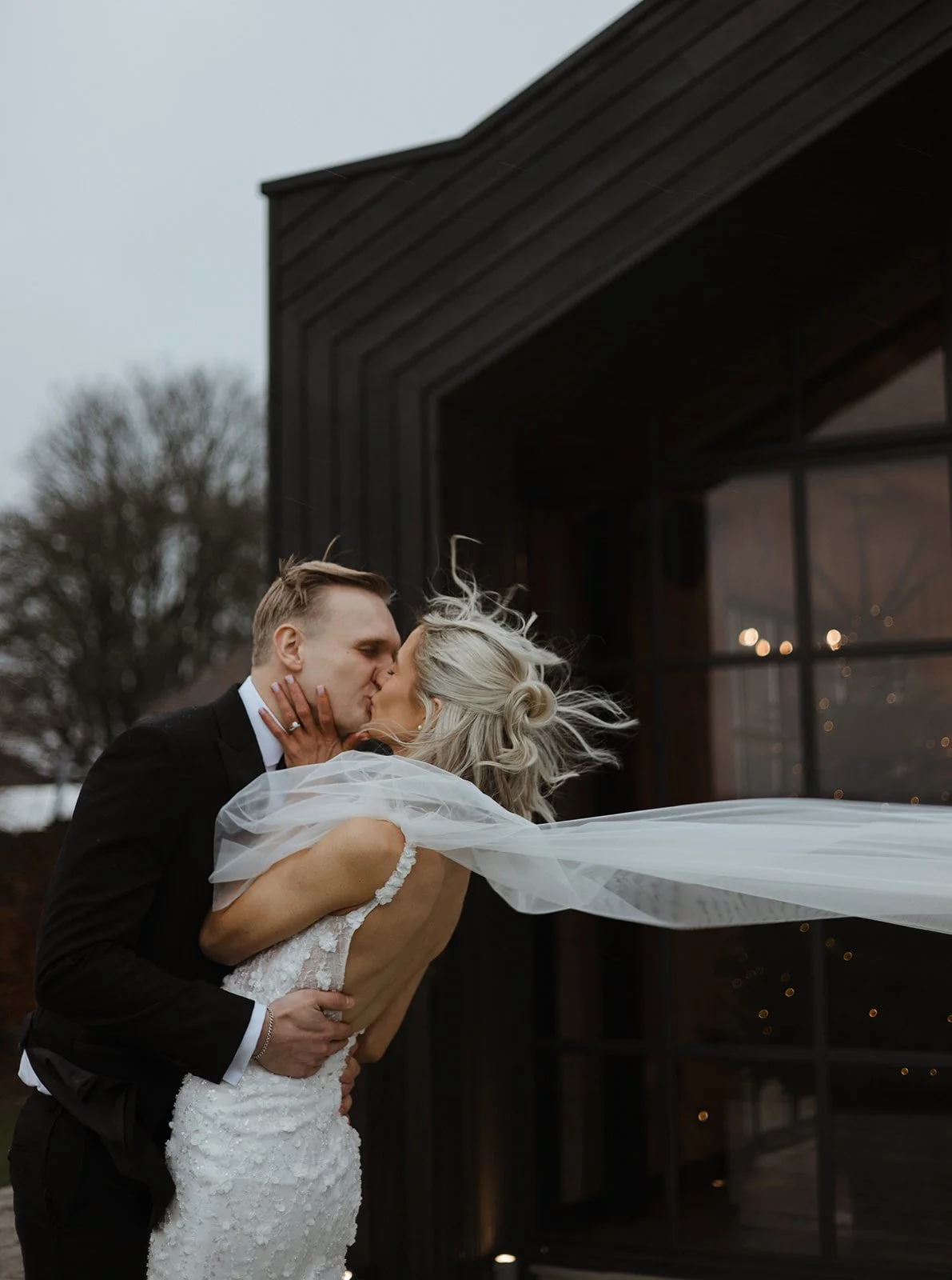A bride and groom kissing outdoors during a wedding, with the bride's veil flowing in the wind and a rustic black building in the background.