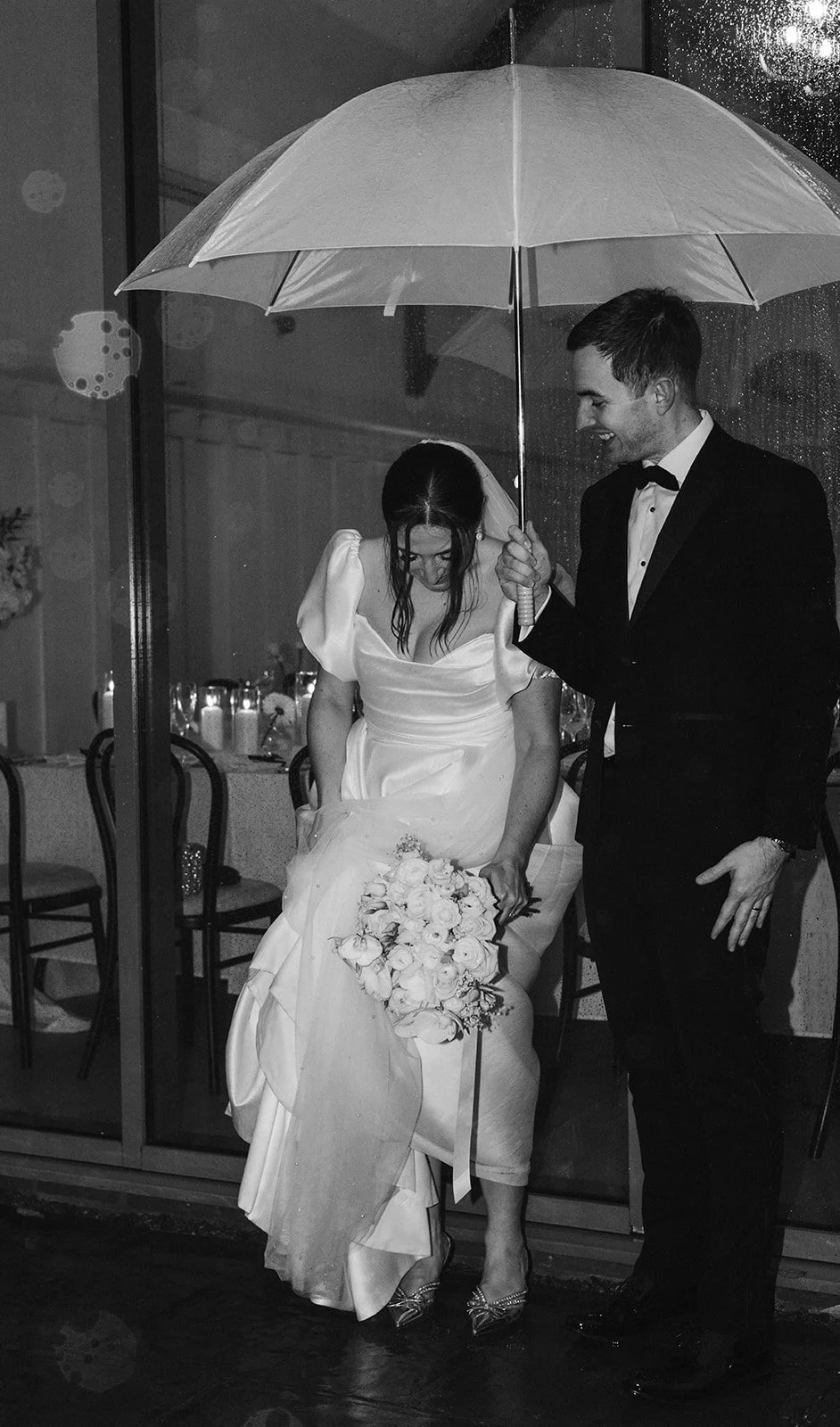 A black and white photo of a bride and groom standing under an umbrella at their wedding reception. The bride is holding a bouquet and looking down, while the groom smiles and holds the umbrella over her.