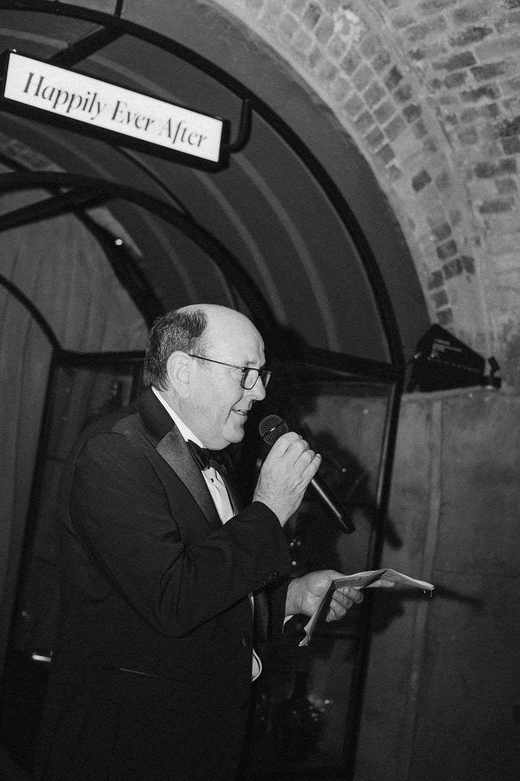 A man in a tuxedo and glasses holding a microphone and reading from a paper, standing in front of a brick archway with a sign that says "Happily Ever After".
