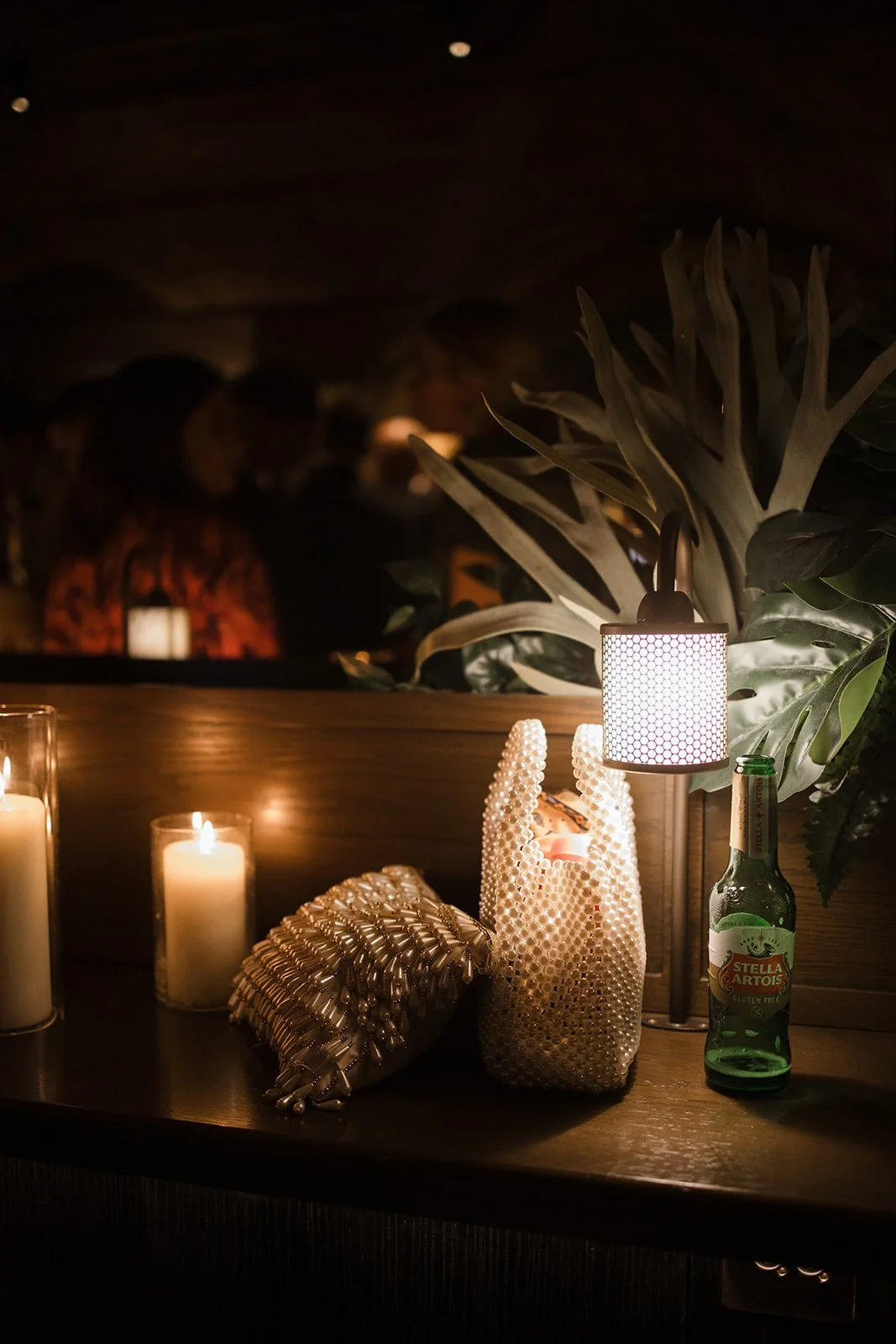 Decorative table setup with candles, a seashell-shaped clutch, a beaded candle holder, a Stella Artois beer bottle, tropical plant, and a hanging lamp, in a dimly lit indoor setting.