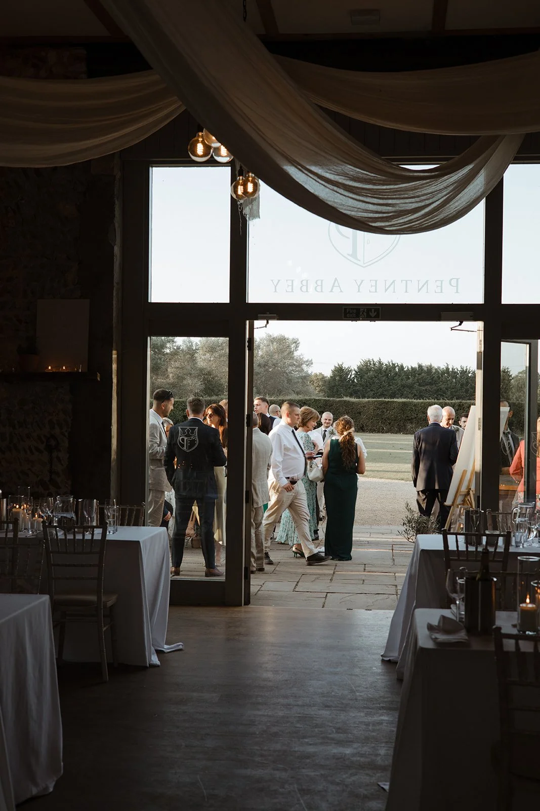People gathered outside a venue, seen through open glass doors, with tables and chairs inside.