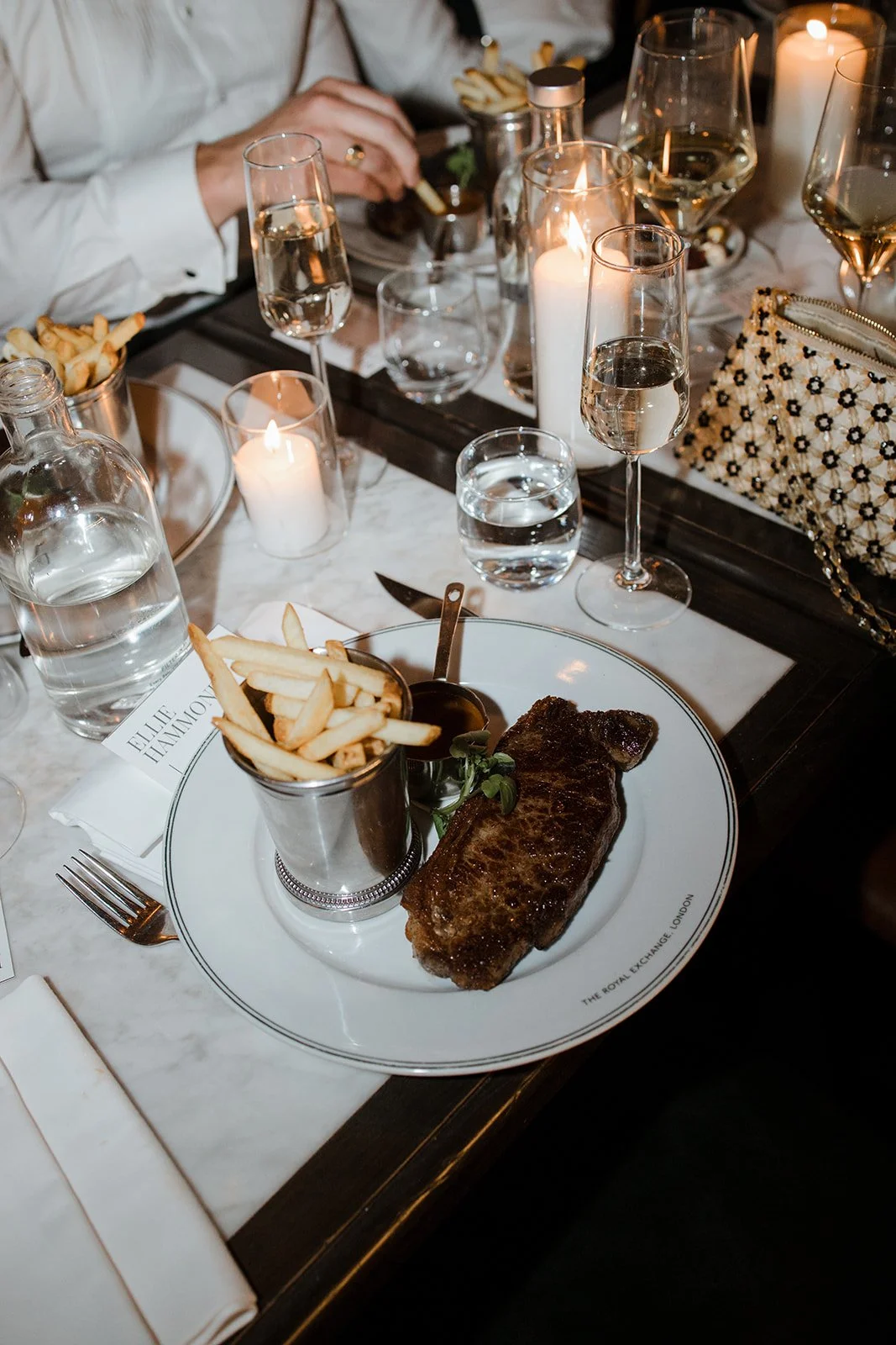 A plated meal with French fries, a beef steak, and sauce, surrounded by wine glasses with white wine, lit candles, and a glass of water, set on a white marble table during a dinner event.
