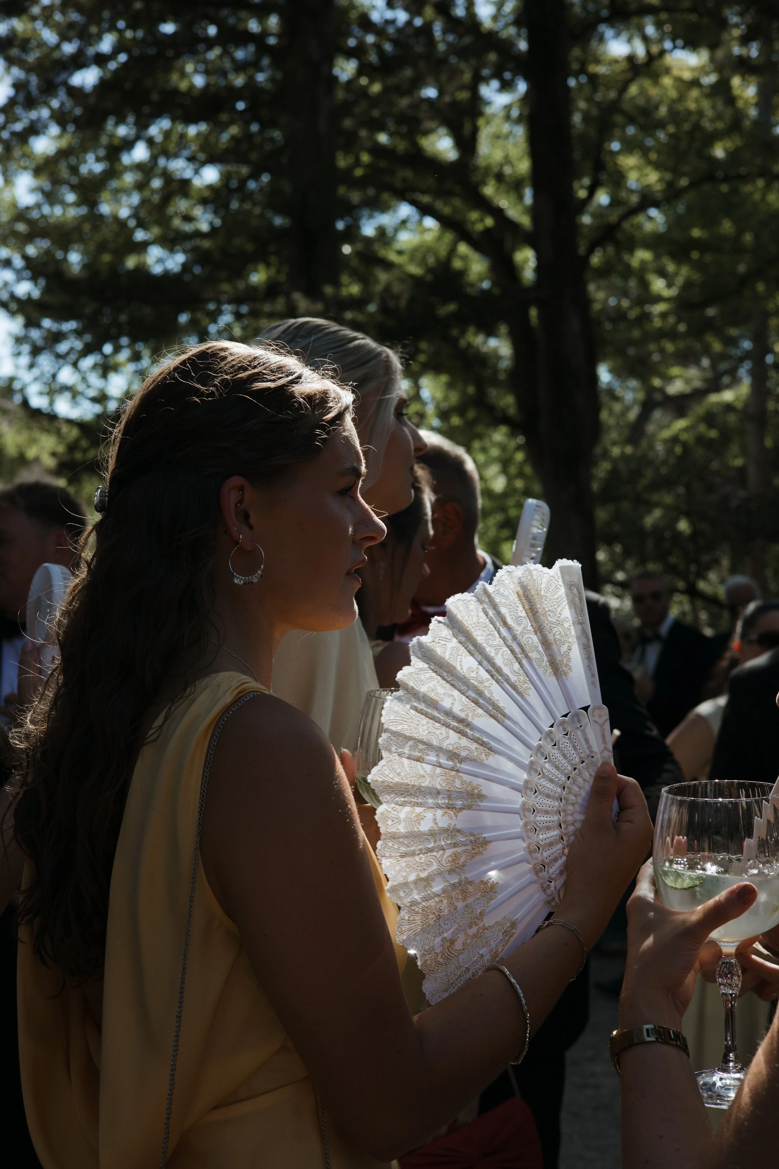 People gathered outdoors during daytime, holding a decorative white fan, with a woman in the foreground wearing a yellow dress and silver earrings, and a glass of white wine in her hand.