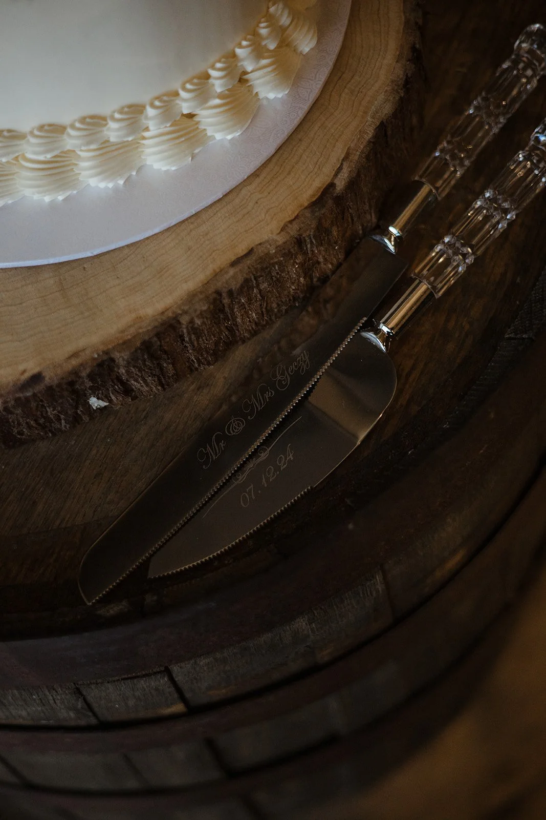 Close-up of a wedding cake with whipped cream piping on top, resting on a wooden surface. Two engraved knives with clear handles are also placed on the surface, with one knife's engraved text reading "Mrs. & Mr. Jelly" and a date, 07.10.24.