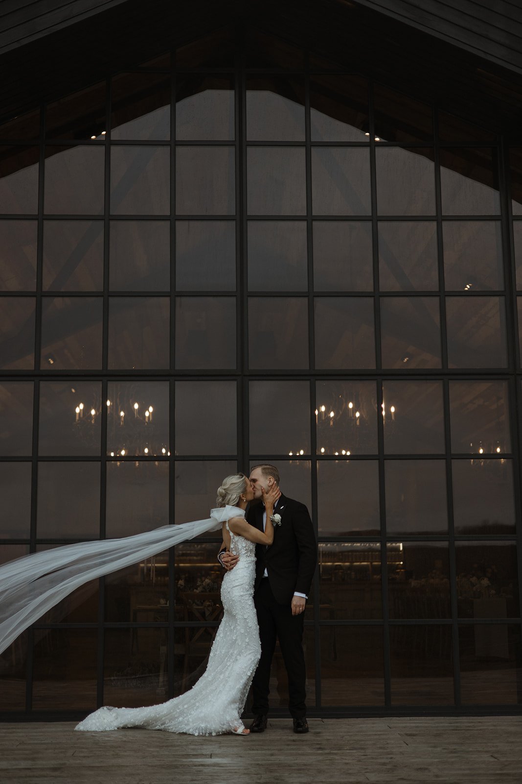A bride and groom kissing in front of a large glass window with chandeliers reflected inside, at sunset or early evening.