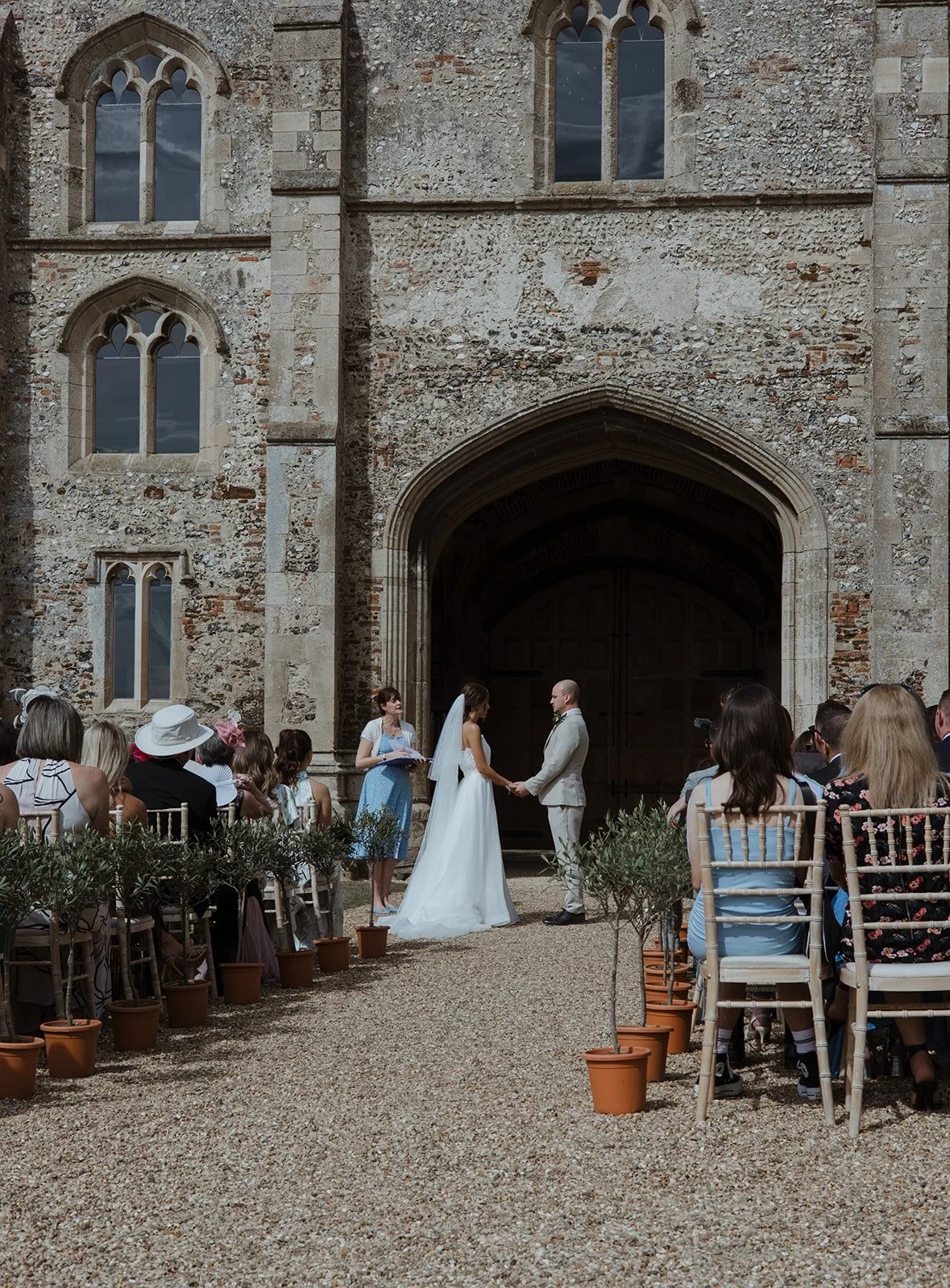A wedding ceremony taking place outside a historic stone church with arched windows. The bride and groom are holding hands at the altar, surrounded by seated guests.