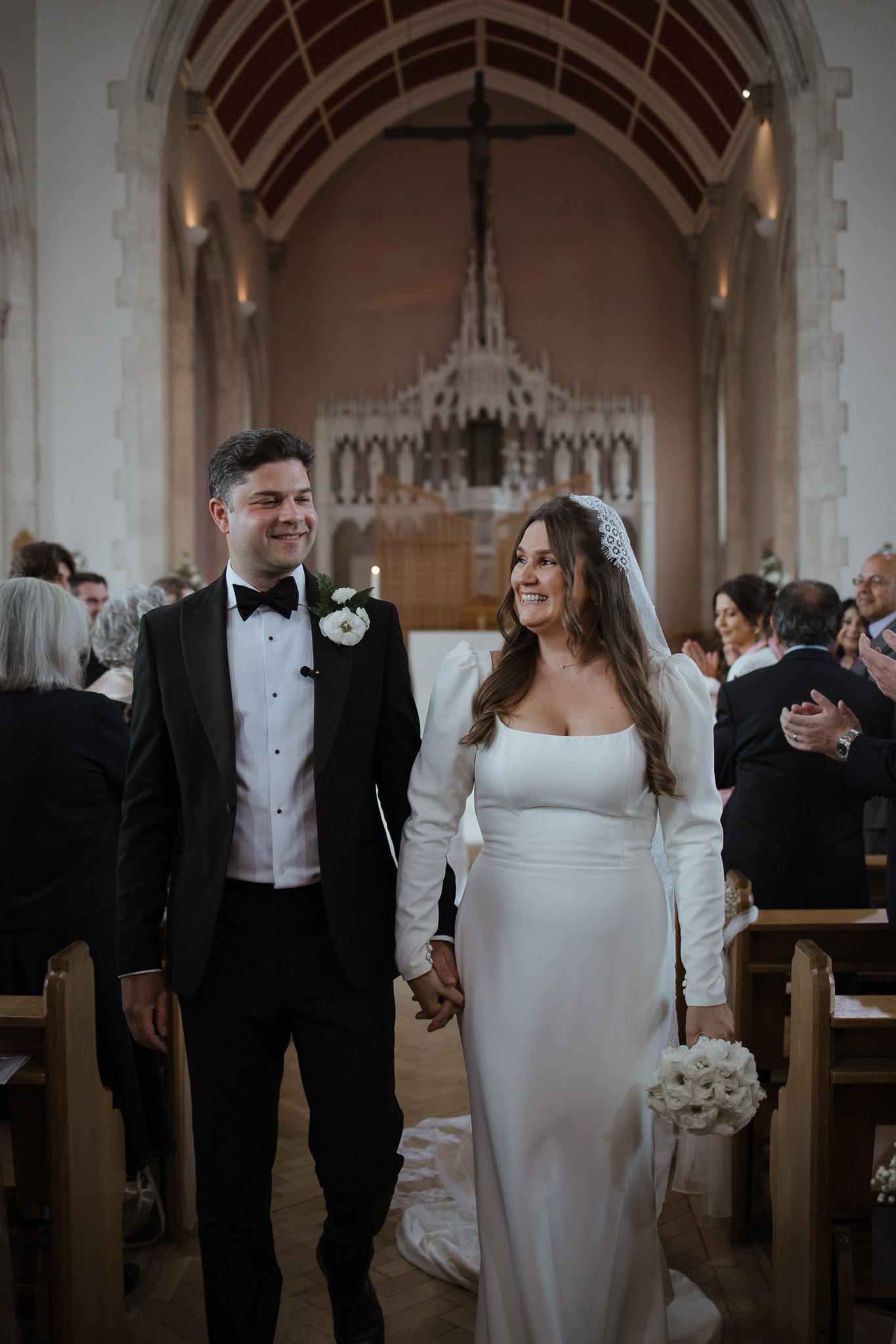 A newly married couple walking down the aisle in a church, smiling and holding hands, with wedding guests clapping and celebrating in the background.