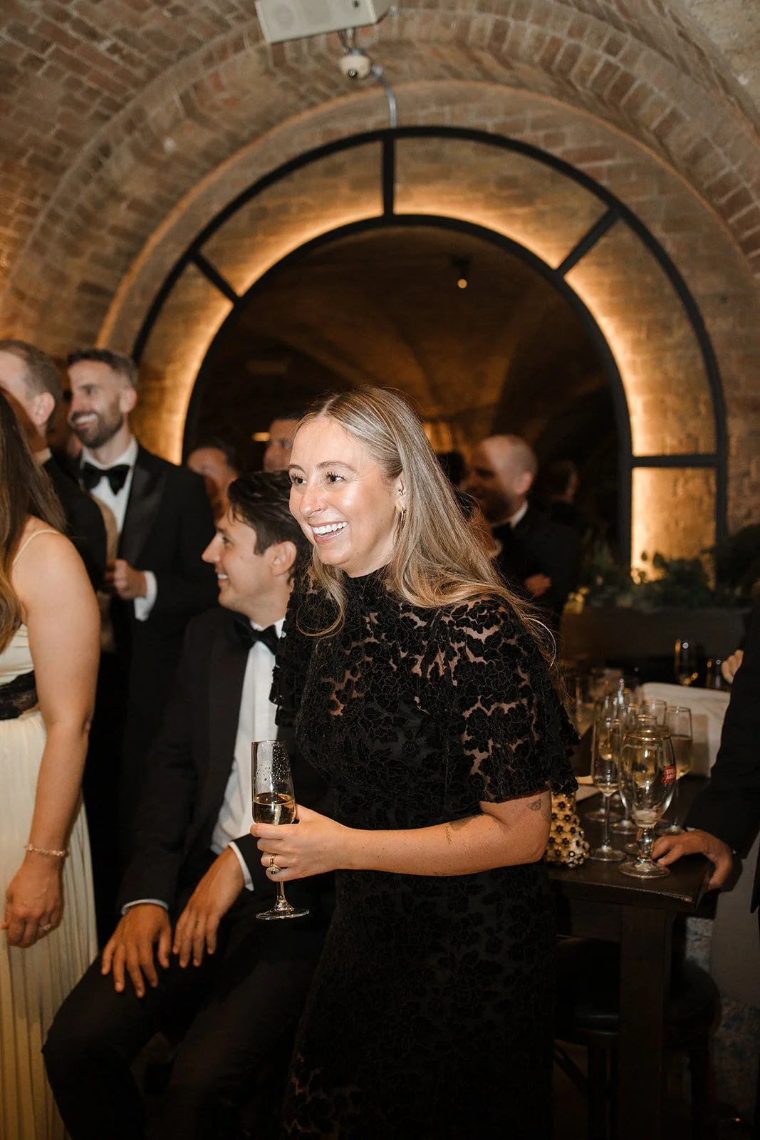 A woman in a black lace dress holding a glass of champagne at a formal gathering or party, with other people dressed in tuxedos and evening gowns in a brick-arched room.