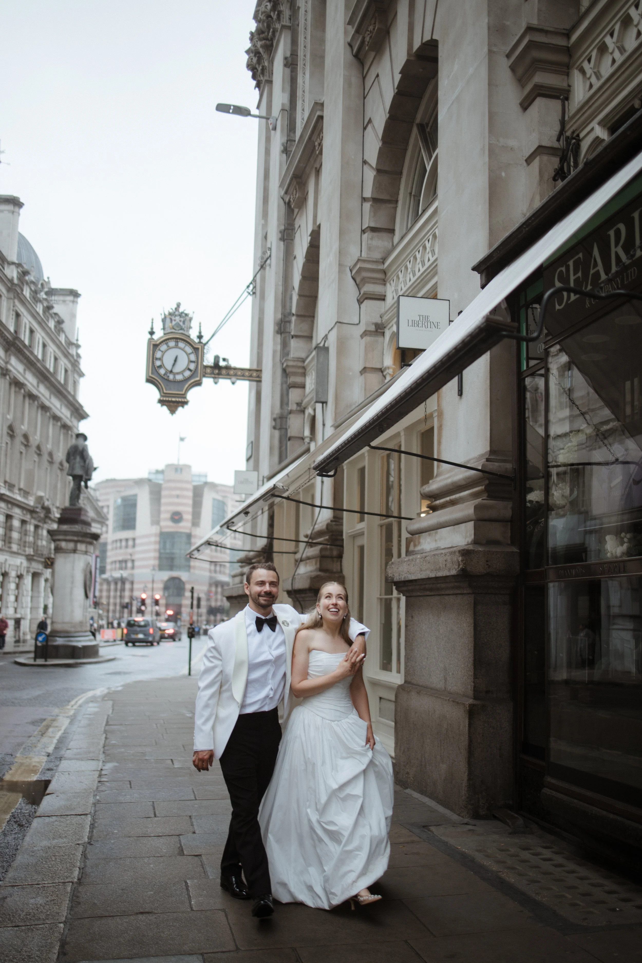 A happy couple dressed in wedding attire walking together on a city sidewalk. The groom is wearing a white tuxedo jacket and black pants, and the bride is in a white wedding gown. They are smiling and holding each other, with tall buildings and a clock hanging on a street sign visible in the background.