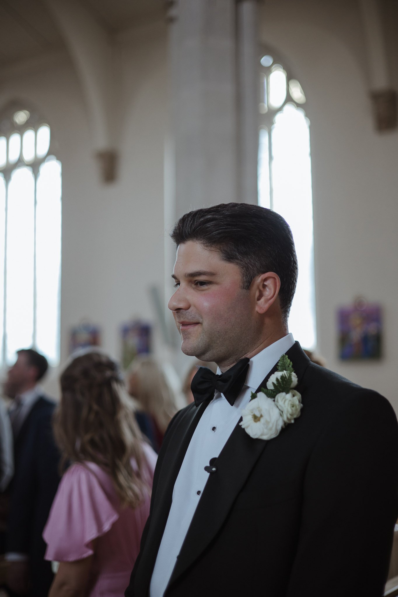 A man in a black tuxedo with a white shirt and black bow tie, standing inside a church, wearing a boutonniere with white flowers and greenery, during a wedding ceremony.