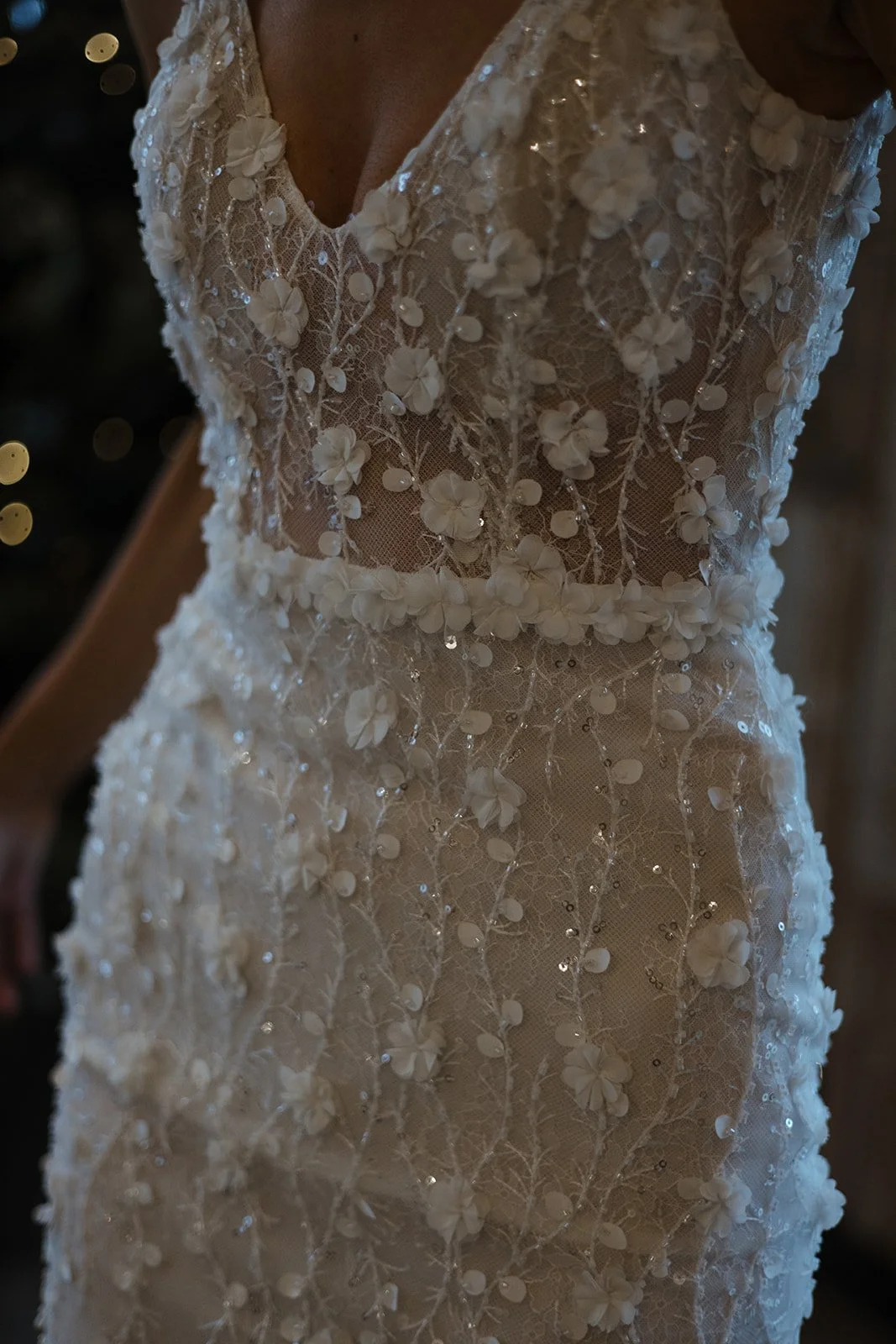 Close-up of a person wearing a richly decorated white wedding dress with 3D floral appliqués, sequins, and lace detailing.