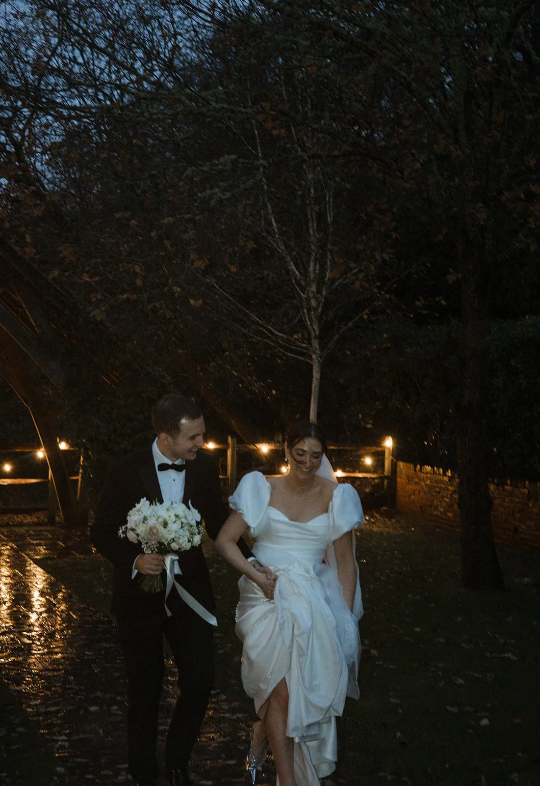 A bride and groom walking outdoors at night, under a tree, with the groom holding a bouquet of white flowers and the bride holding her dress.