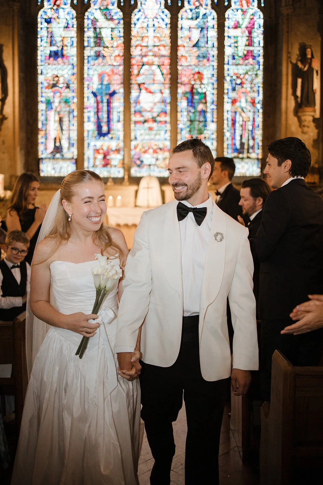 Bride and groom smiling and holding hands inside a church with stained glass windows in the background, surrounded by guests.