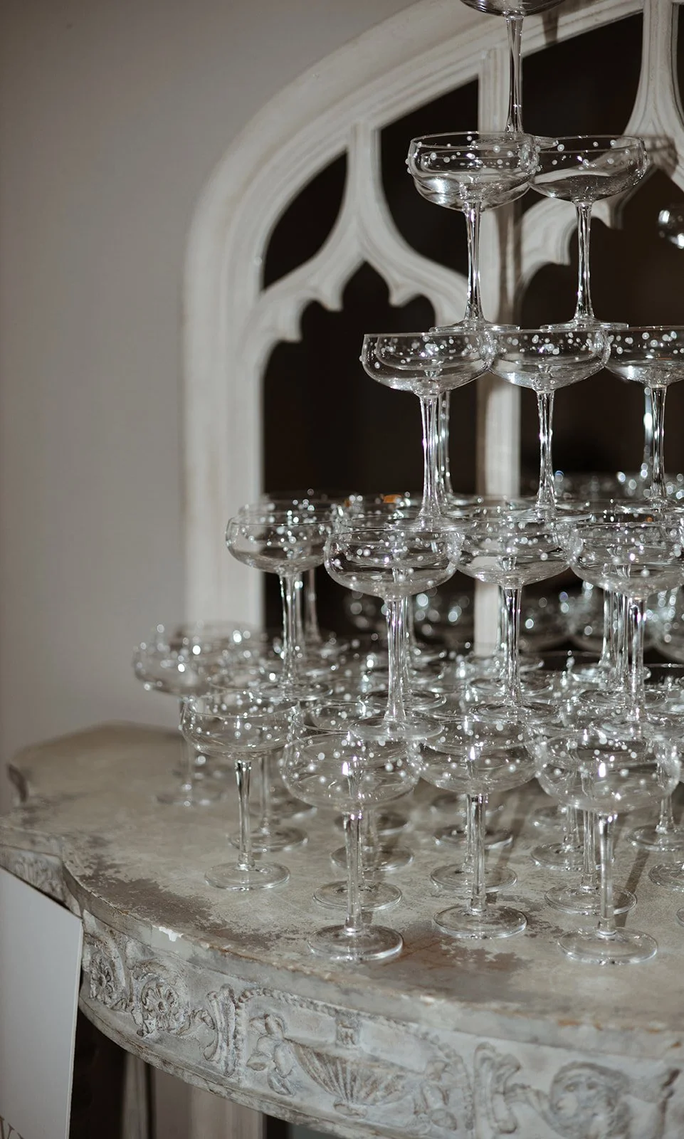 A pyramid of empty champagne coupes stacked on a vintage table with ornate carvings, with a decorative mirror in the background.