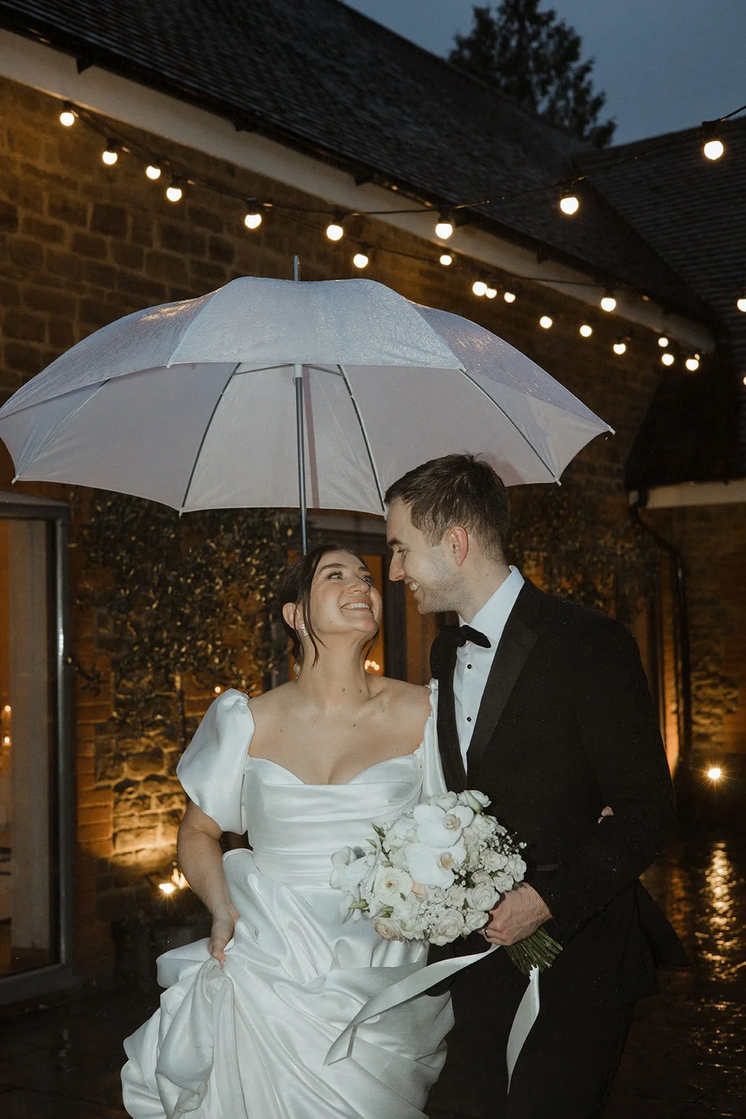 A bride and groom share a joyful moment under an umbrella during their wedding celebration at night, with string lights overhead and a rustic building in the background.