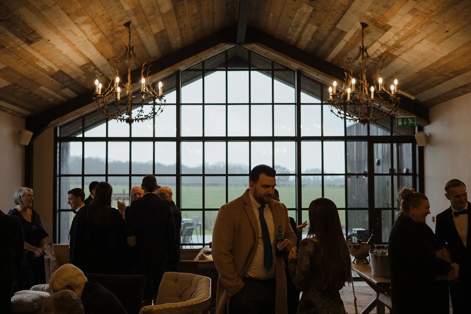 People socializing at an indoor event with large industrial-style windows and chandeliers, with a view of a green field outside.