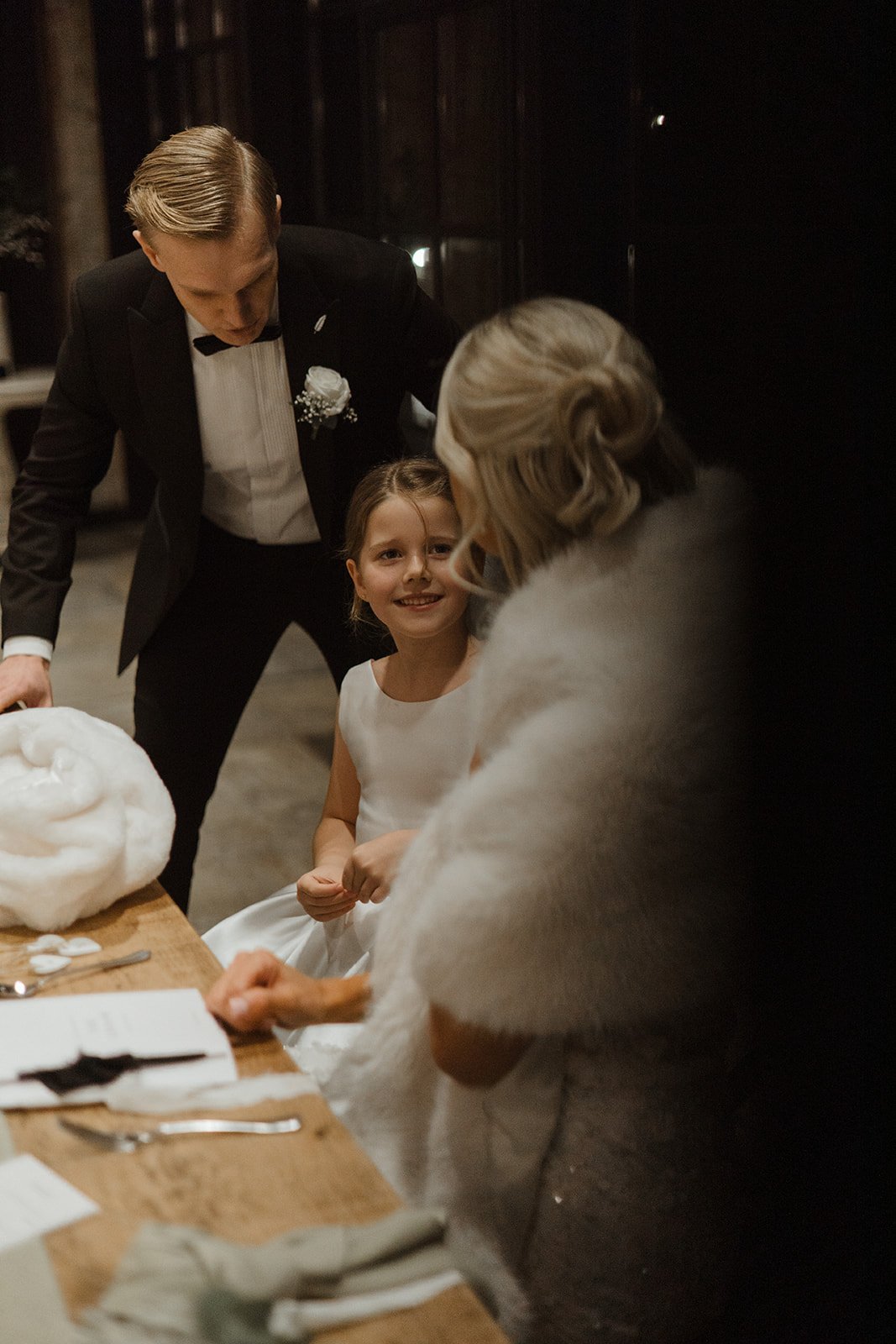 A young girl in a white dress interactions with an elderly woman dressed in a light-colored coat, while a man in a tuxedo looks on, during a special event or celebration indoors.