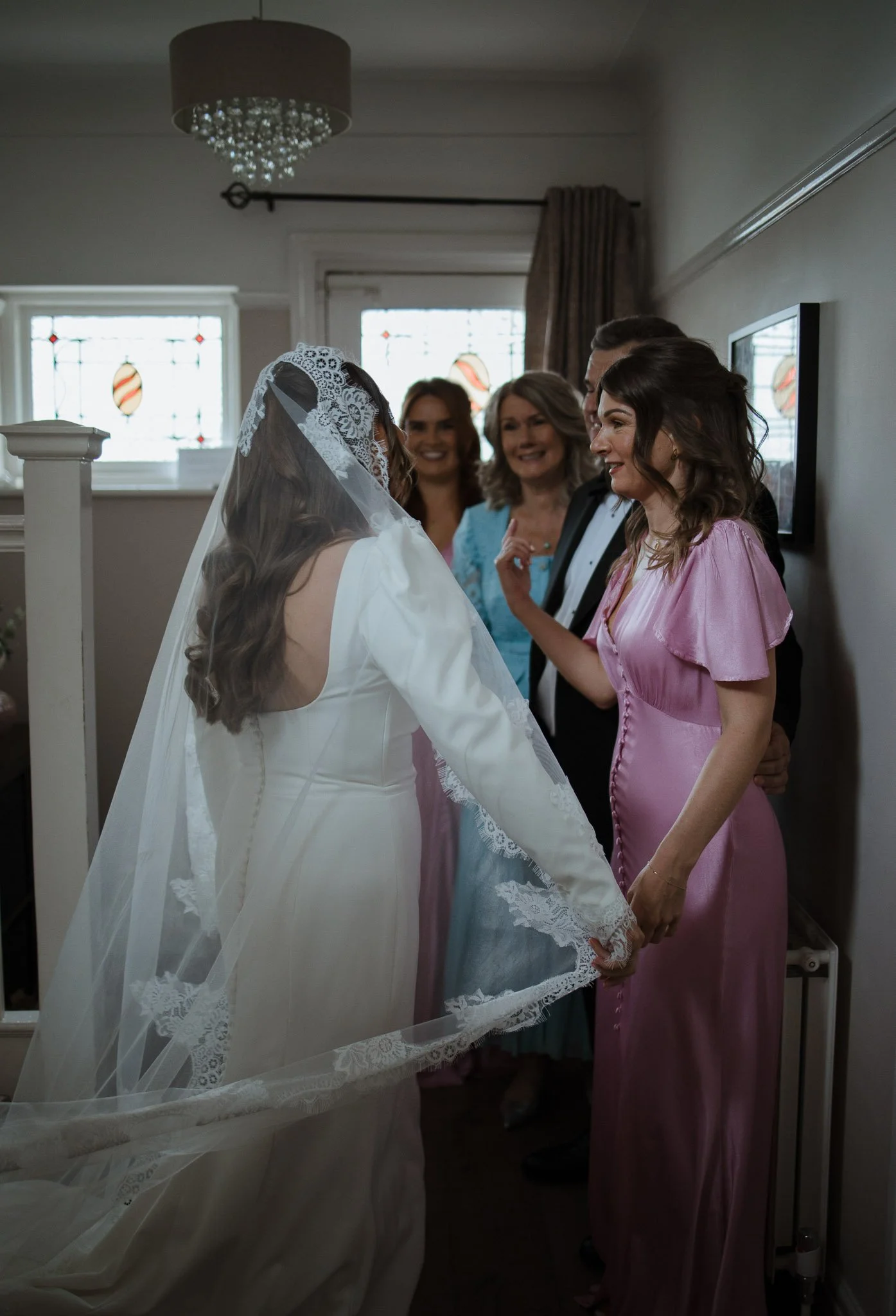 A bride in a white dress and veil holding hands with a woman in a pink dress during a wedding preparation, with three smiling women in the background.