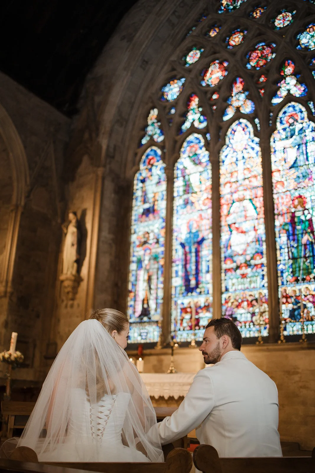 A bride and groom during their wedding ceremony inside a church, with a large stained glass window in the background.