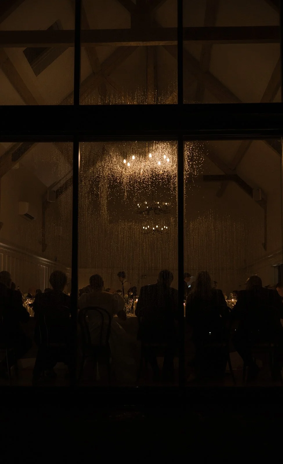 A dark room seen through a large window with raindrops on the glass, showing a chandelier and a group of people seated at tables, likely at a dinner event.
