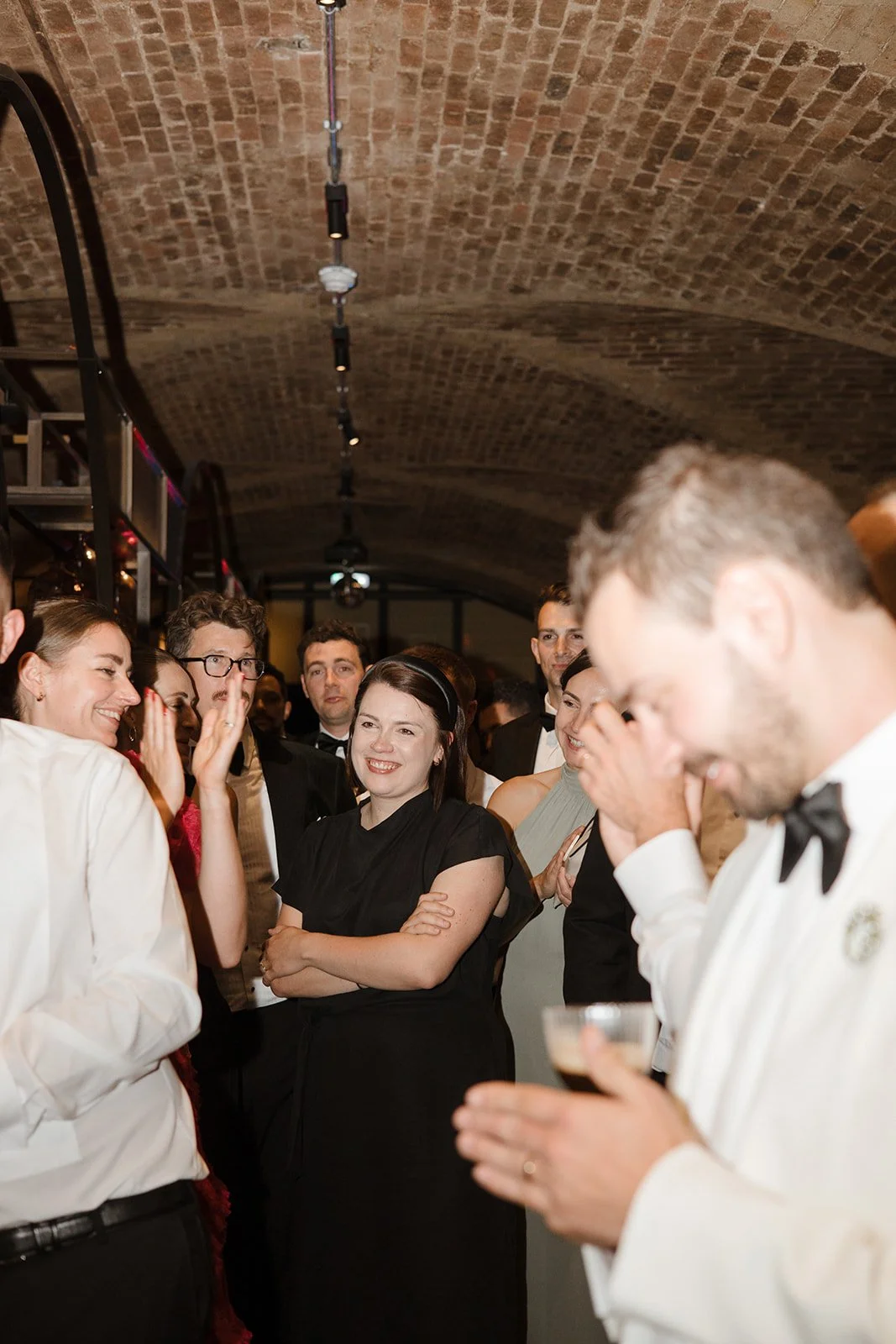 Group of people at a social gathering in an indoor venue with a brick arch ceiling. They are dressed in formal attire, with some wearing tuxedos and others in elegant dresses. The woman in the center is smiling, while others are engaged in conversati
