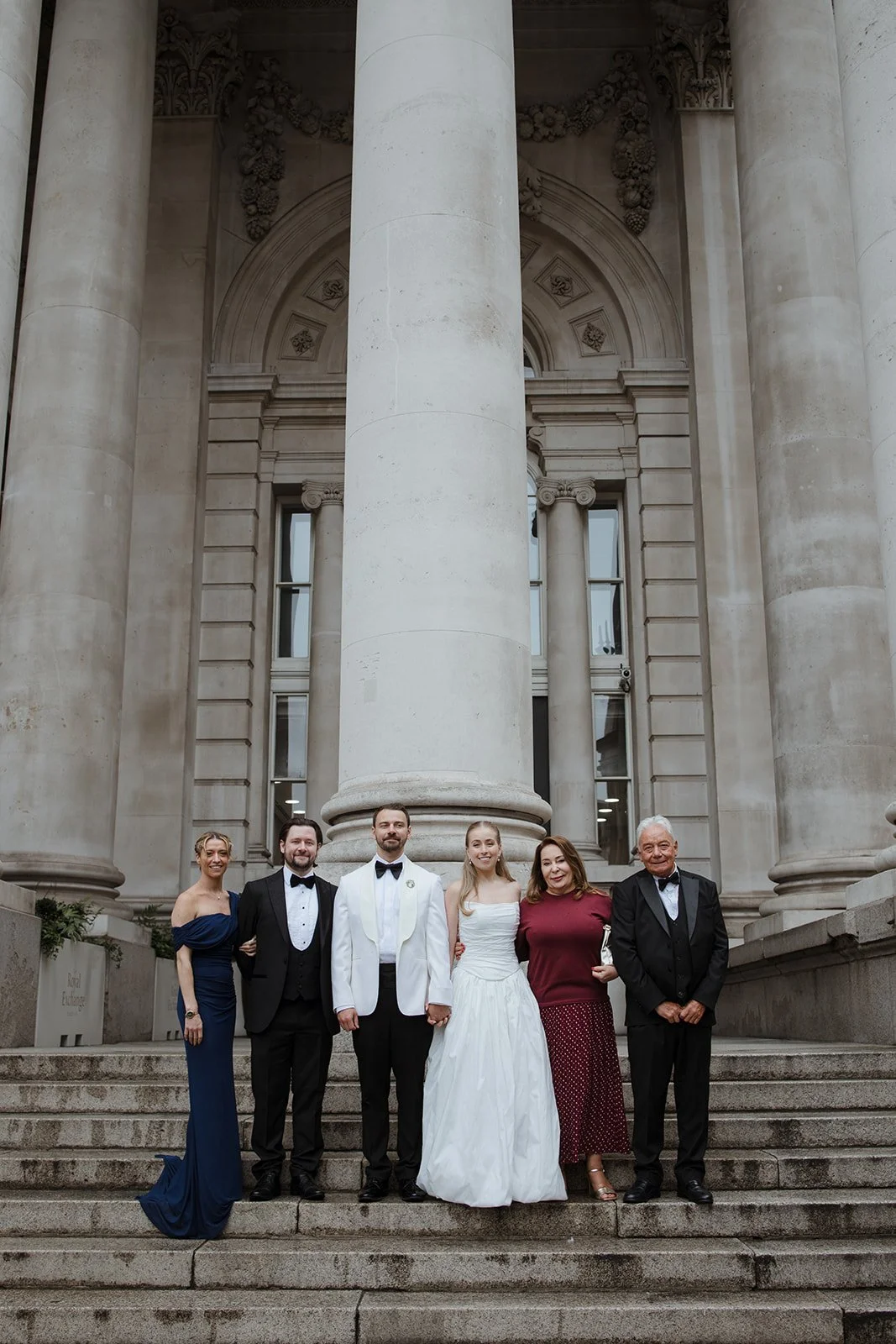 A group of six people standing on steps in front of a building with large stone columns. One woman is in a dark blue gown, and the bride is in a white wedding dress. The men are in tuxedos, and the others are dressed in formal attire.